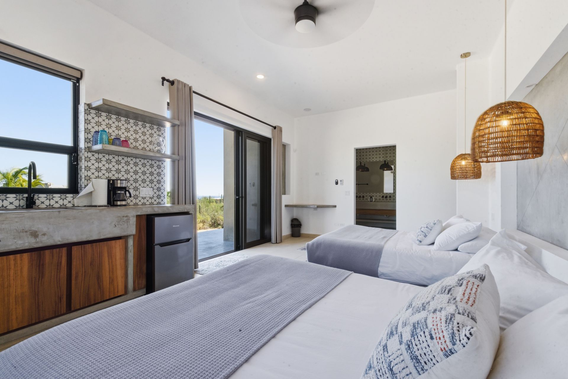 Bright hotel room with two beds, kitchenette, and balcony access.