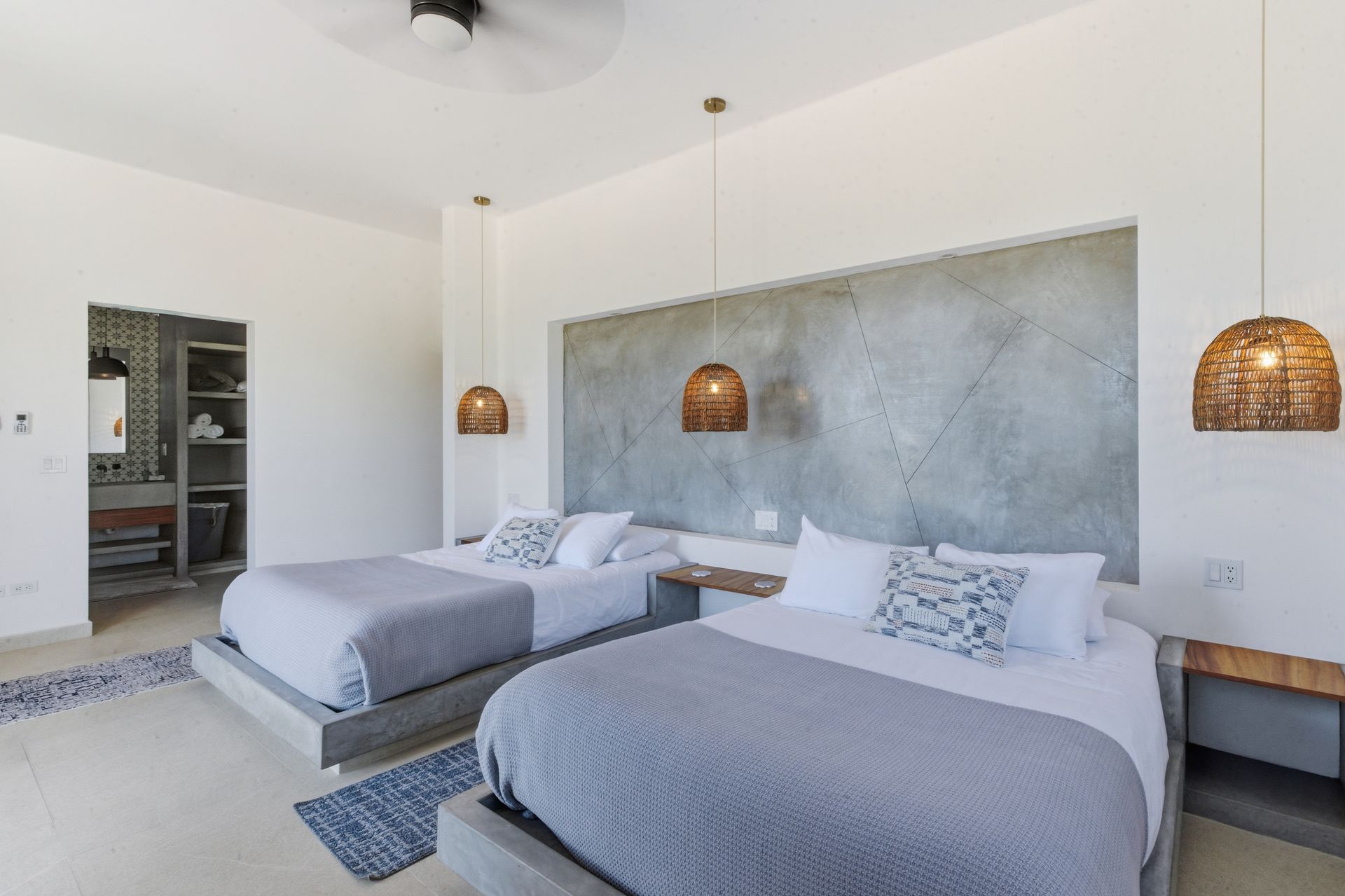 Bedroom with two beds, gray blankets, woven pendant lights, and concrete headboard. A bathroom is in the background.