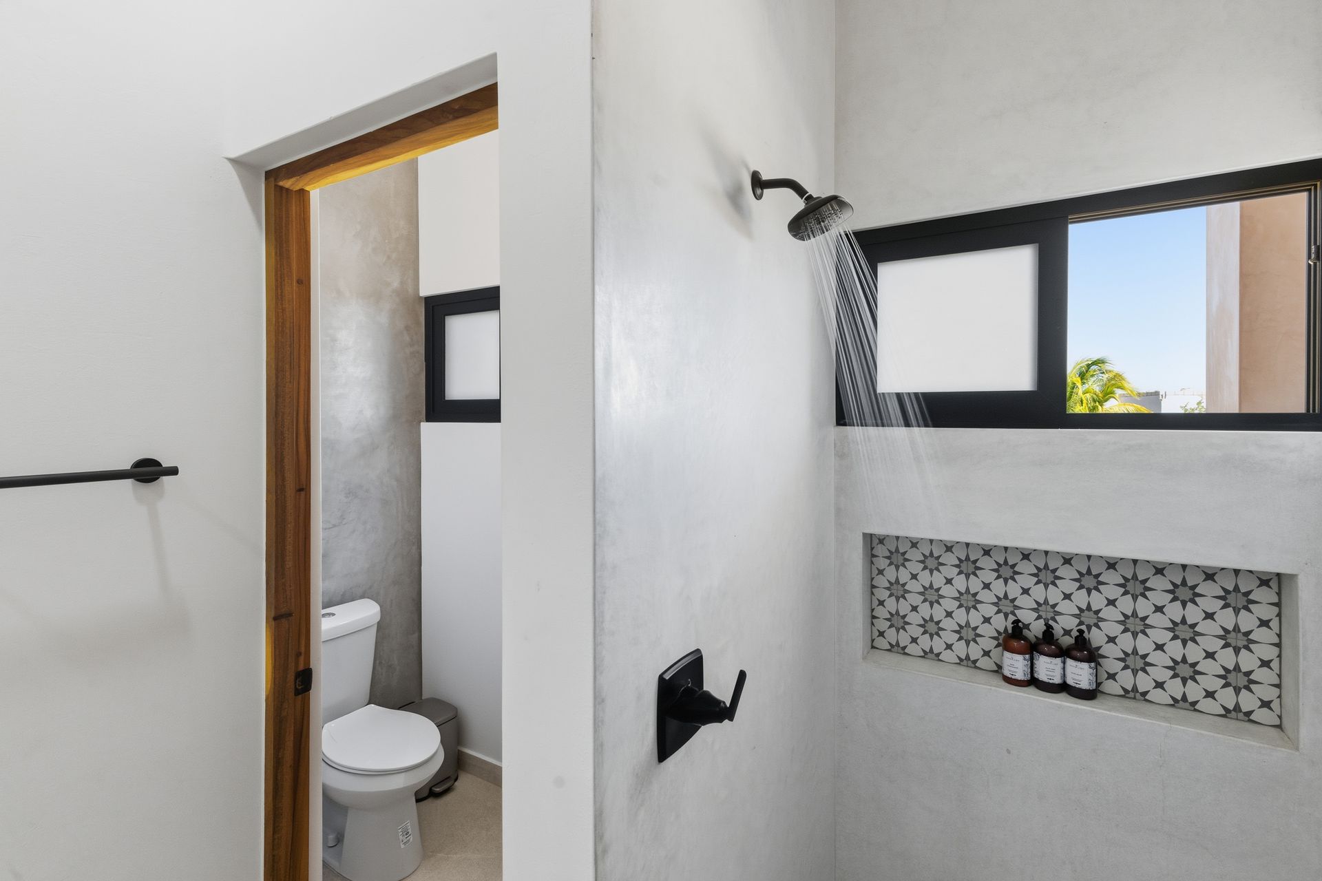 Bathroom with shower, toilet, window. White walls, wood door, black fixtures, patterned tile, natural light.