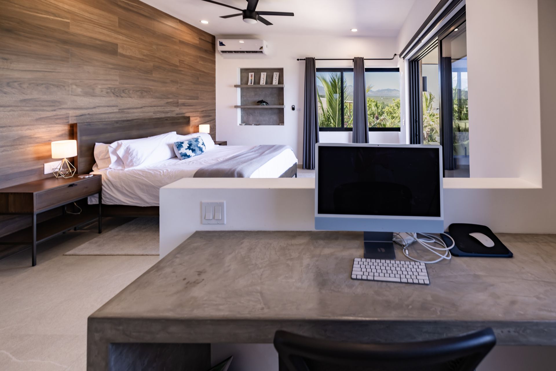 Bedroom with wooden accent wall, king-size bed, desk with computer and phone, and sliding glass doors.