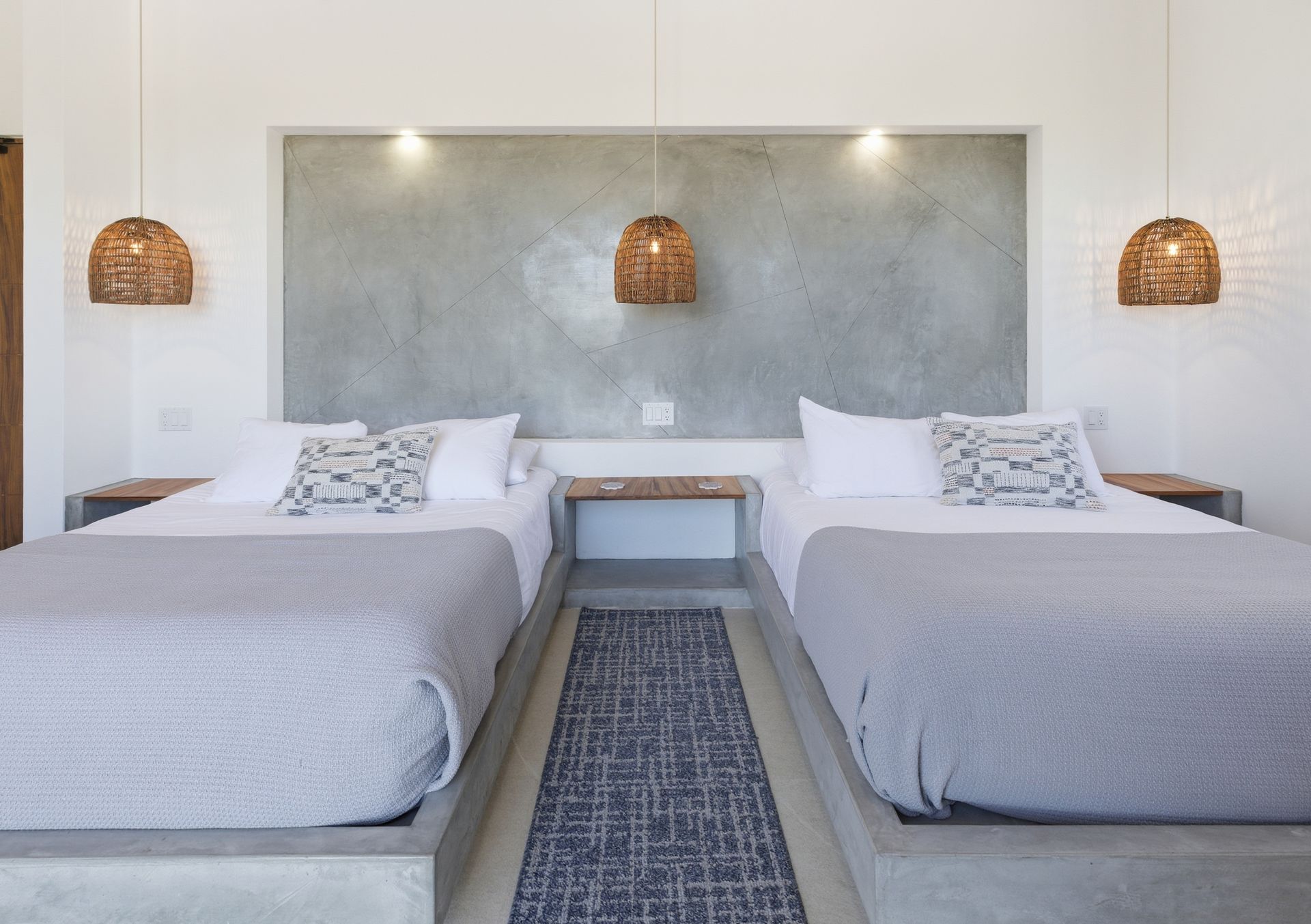 Two beds in a minimalist bedroom with concrete accents, woven lamps, and gray bedding.