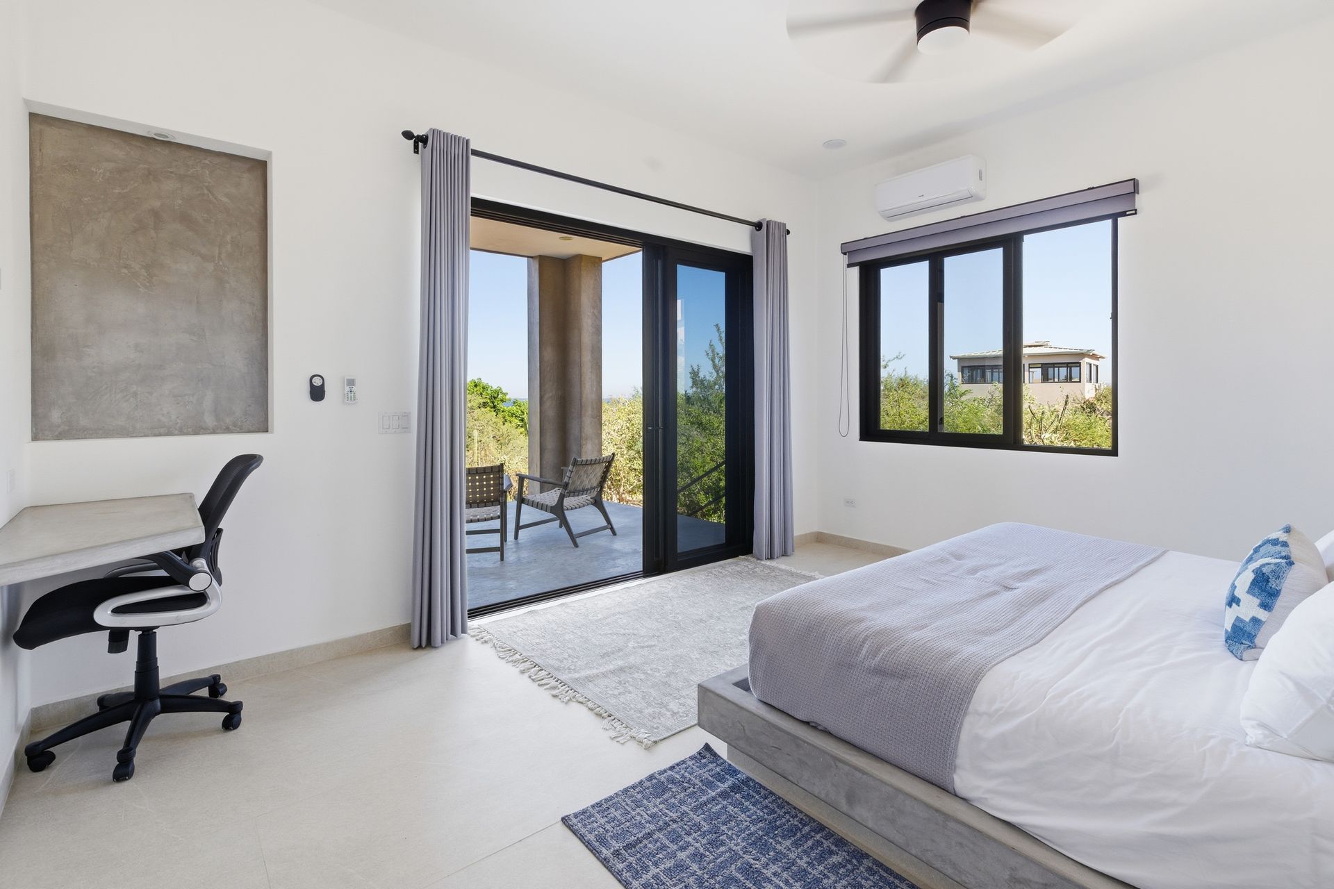 Bedroom with bed, desk, and balcony access. White walls, sliding glass doors, neutral color scheme.