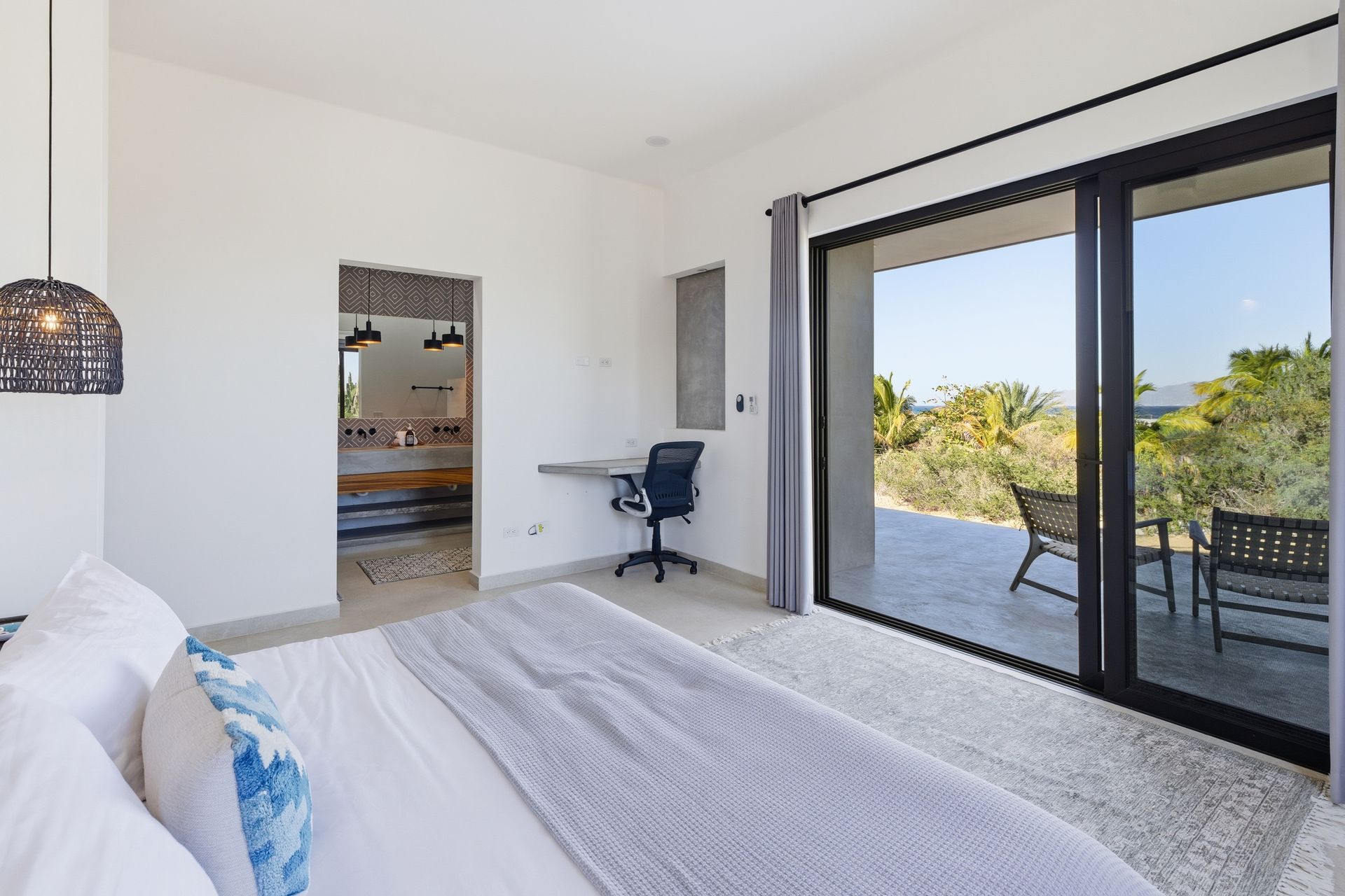 Bedroom with open patio doors; bed, desk, and outdoor seating visible. Bright natural light.