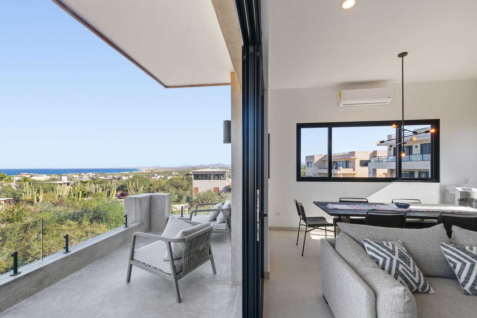 Bright, modern interior with balcony overlooking a coastal landscape. Grey furniture, sliding glass doors.