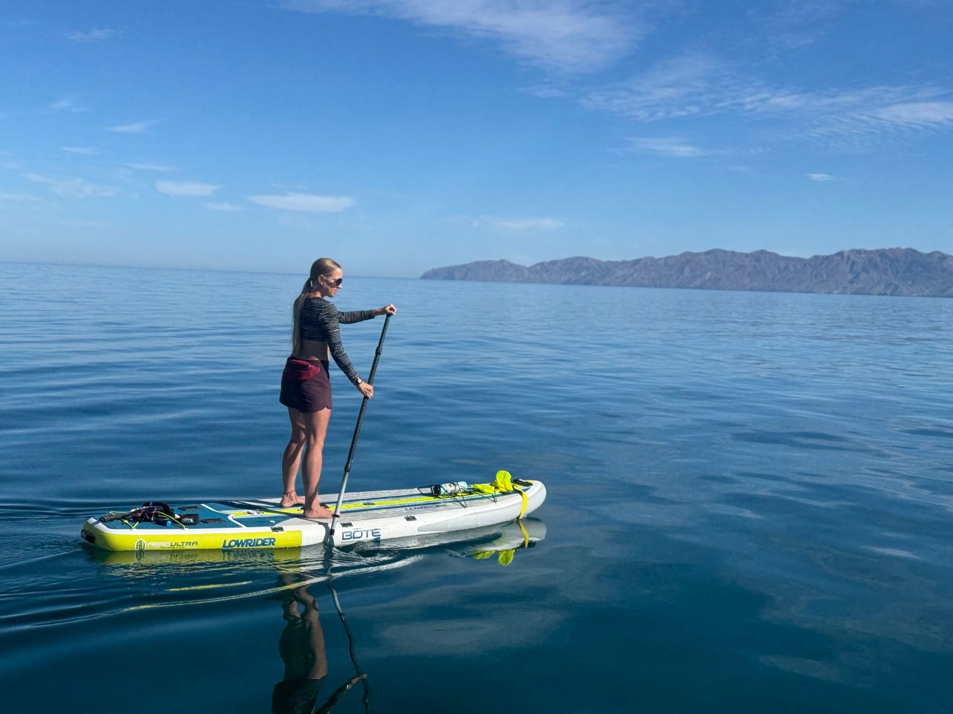 Woman paddleboarding on calm, blue water towards distant land under a clear sky.