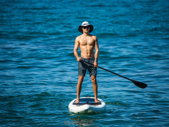 Man paddleboarding on a blue ocean, wearing hat and sunglasses, holding a paddle.