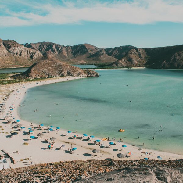 Scenic beach with turquoise water, white sand, and blue umbrellas. Mountains in the background, sunny sky.
