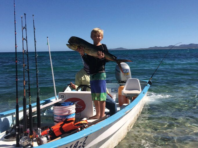 Boy on a small boat holding up a large fish, blue water and coastline in background.