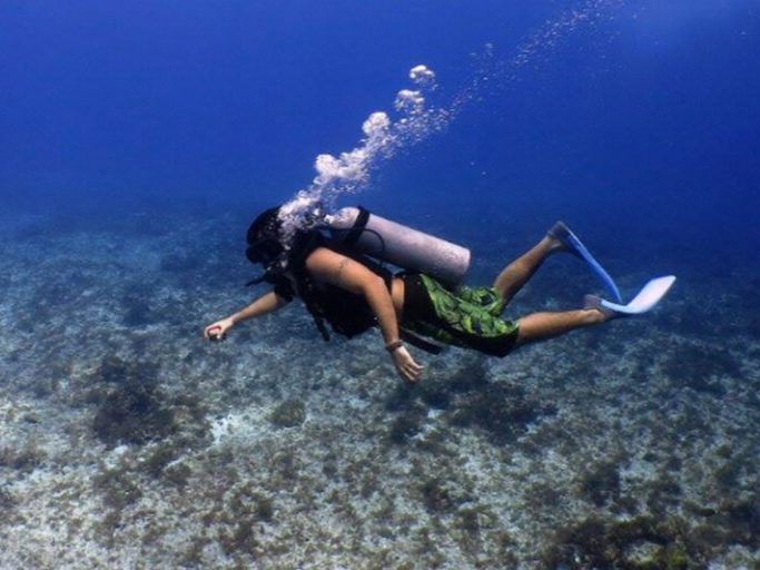 Scuba diver swimming underwater, exhaling bubbles; bright blue water, coral reef.