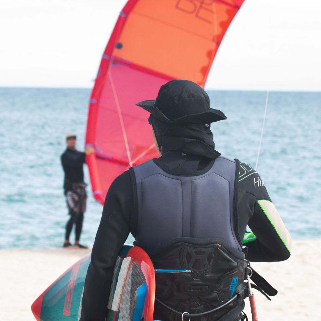 Person in wetsuit holding a surfboard, facing the ocean, kite in the wind. Another person and kite in the background.