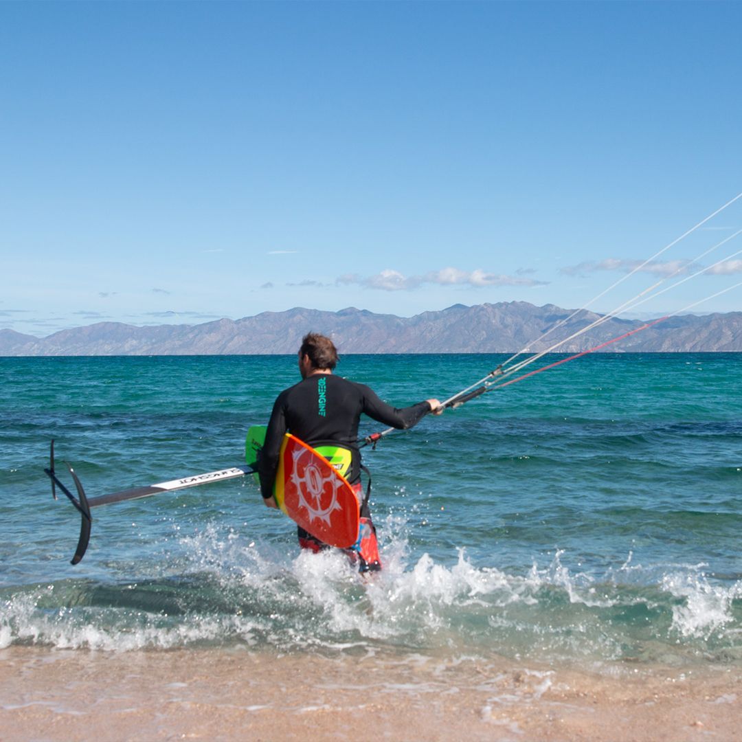 Man in wetsuit kiteboarding in turquoise water, holding lines and foil board near a beach.