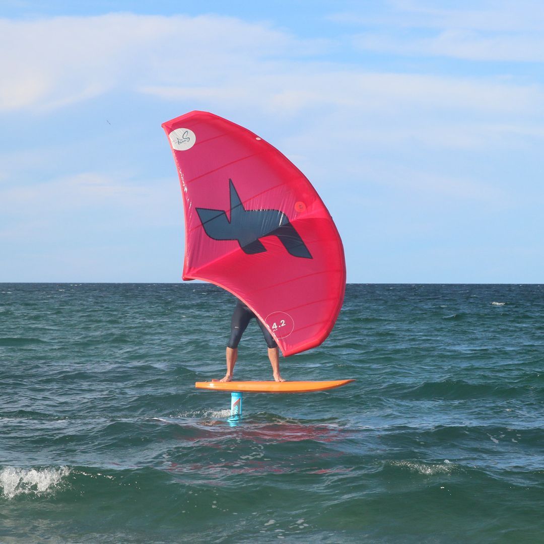 Person on hydrofoil board, holding a red wing sail, gliding on water. Blue sky and ocean in the background.