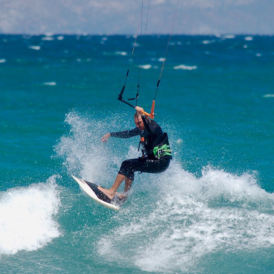 Kitesurfer on a white board carving through a wave, ocean backdrop with a blue kite overhead.