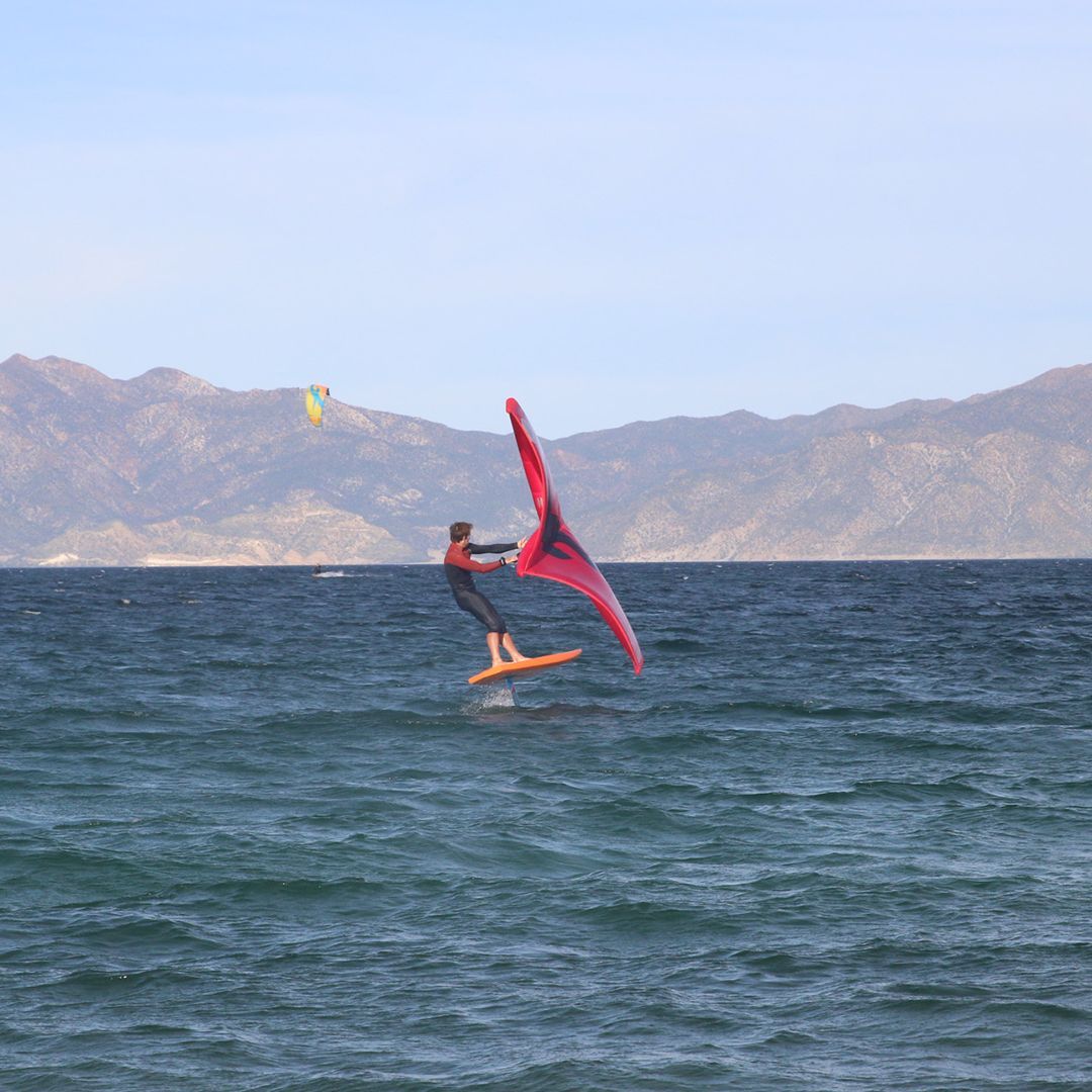 Person wing foiling on a board, lifted above ocean surface, holding red wing. Mountains in background.