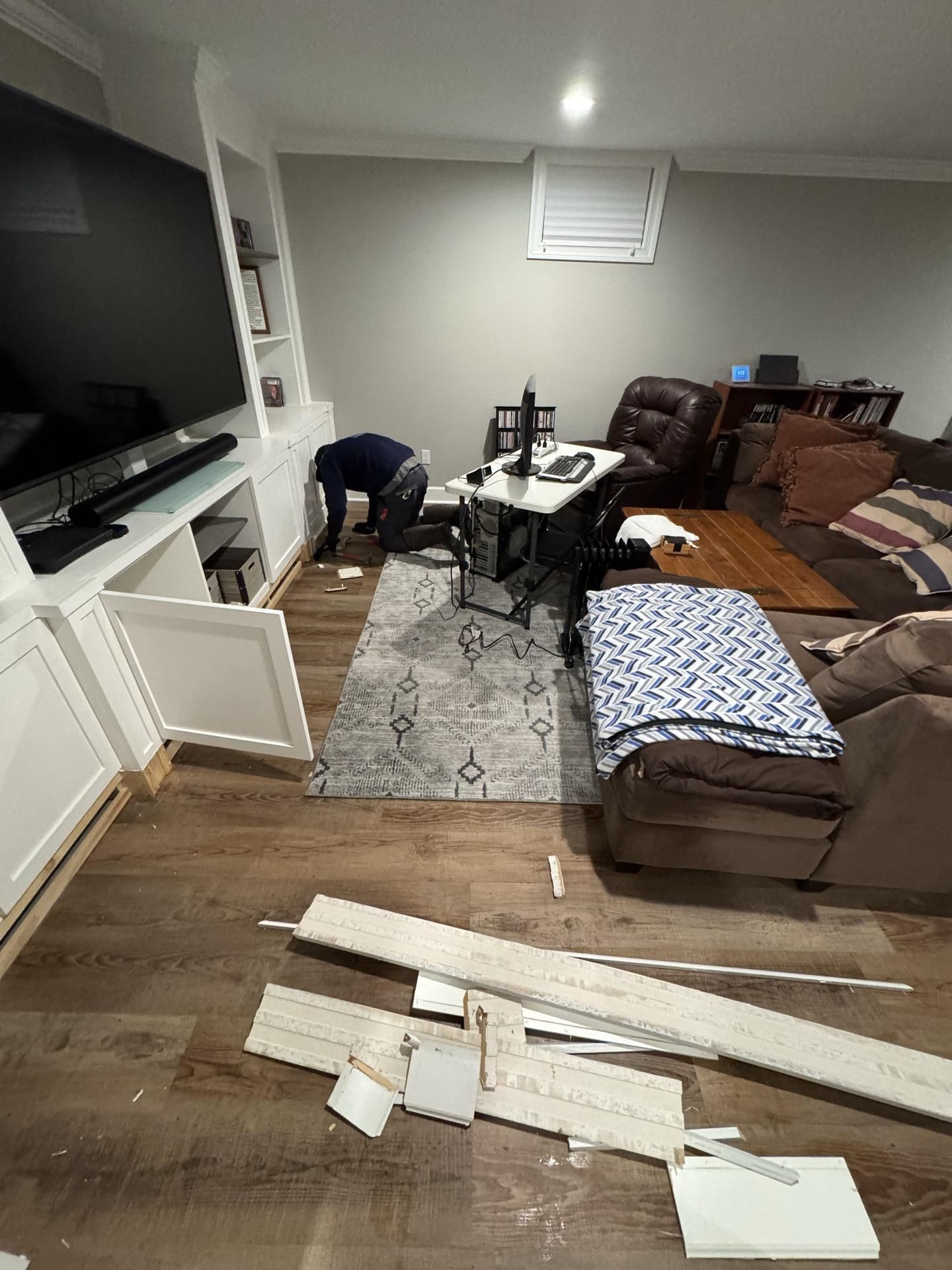 A person kneels to work on a white built-in cabinet in a living room, with scattered construction materials on the floor.