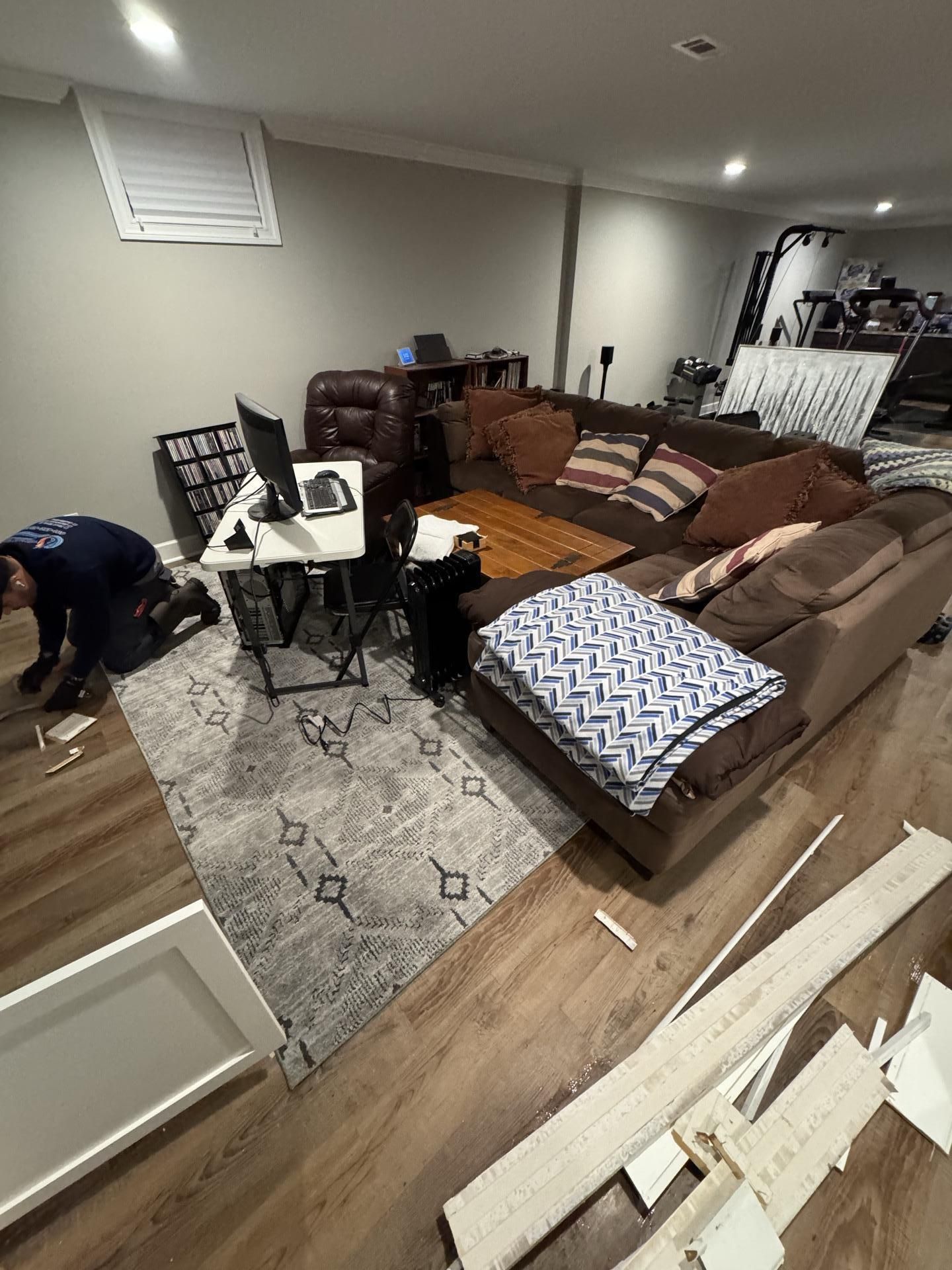 A person works on a basement floor next to a white desk, a brown sofa, and scattered construction materials.