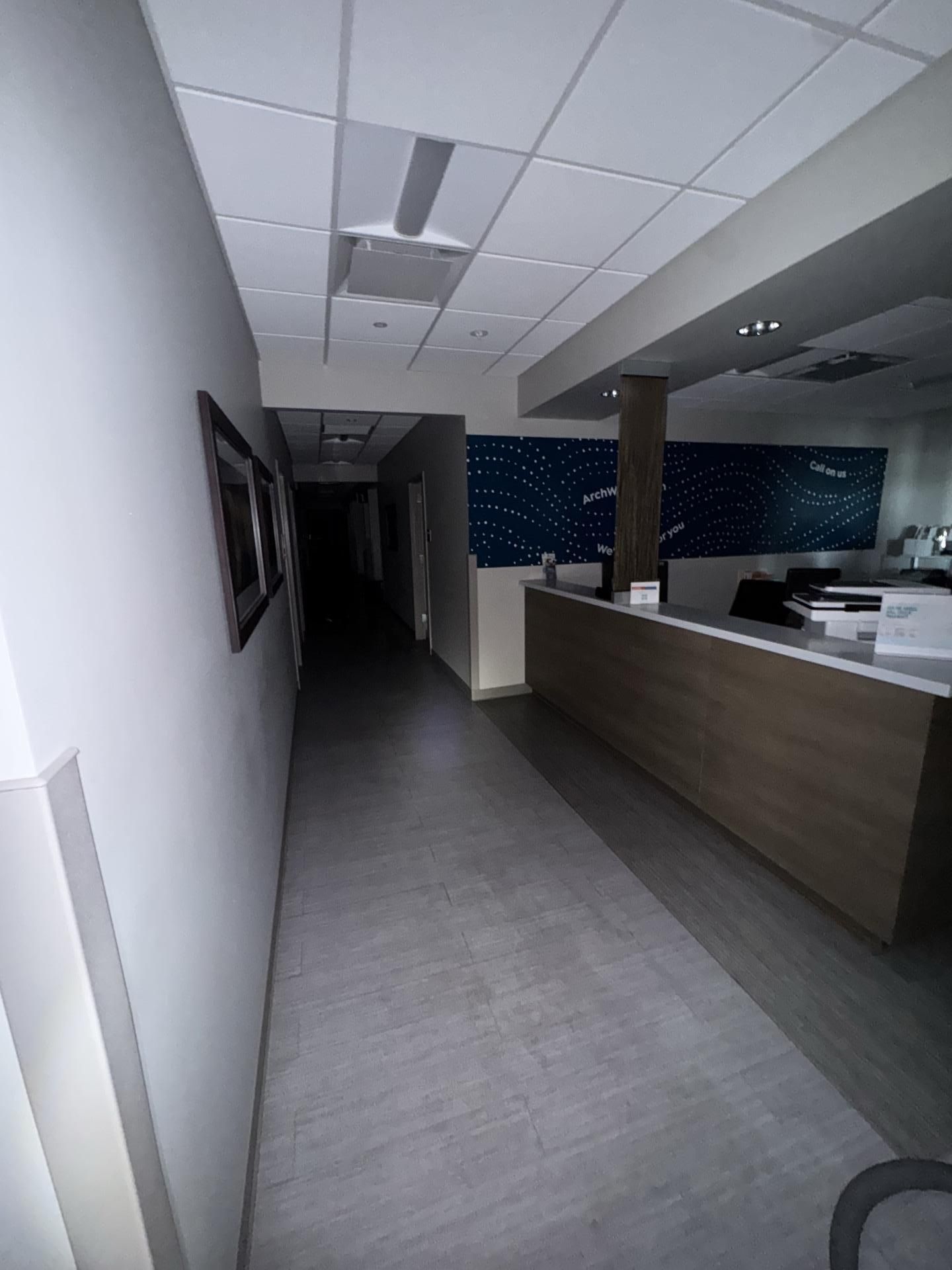 An indoor hallway leads to a dark corridor, with a reception desk on the right featuring a blue wall display.