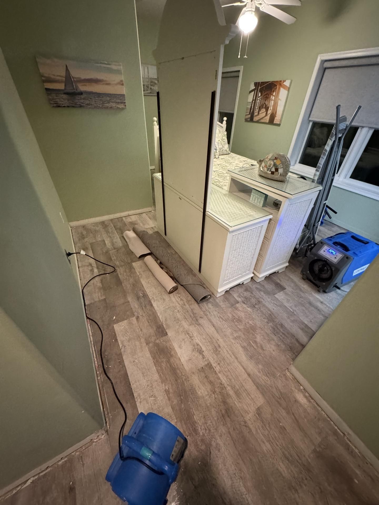 A bedroom under restoration with two blue air movers on the floor, rolled-up rug remnants, and furniture pushed to the side.