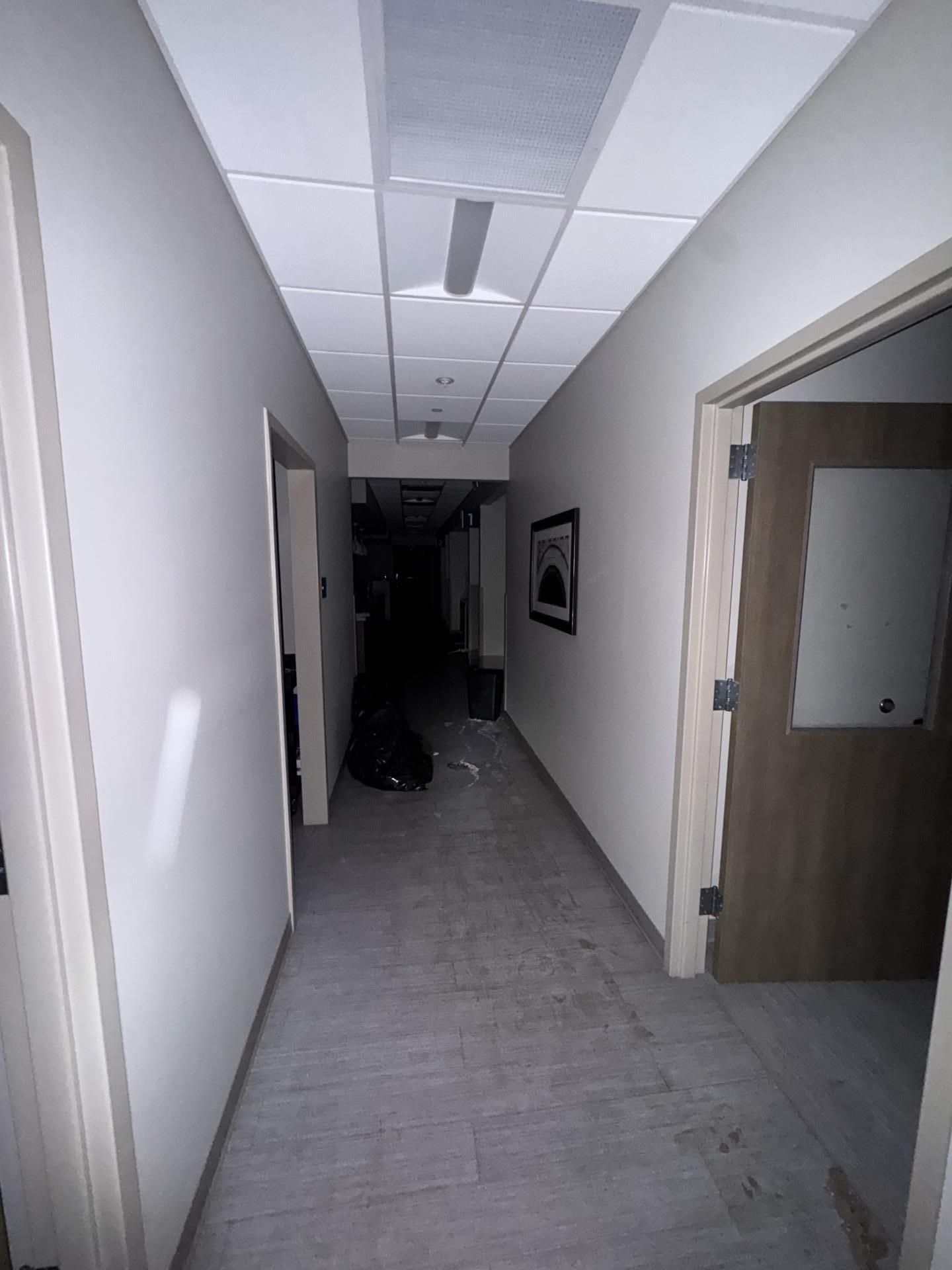 A dimly lit office hallway with white walls, tiled ceilings, and a light-colored floor, leading toward a dark area.