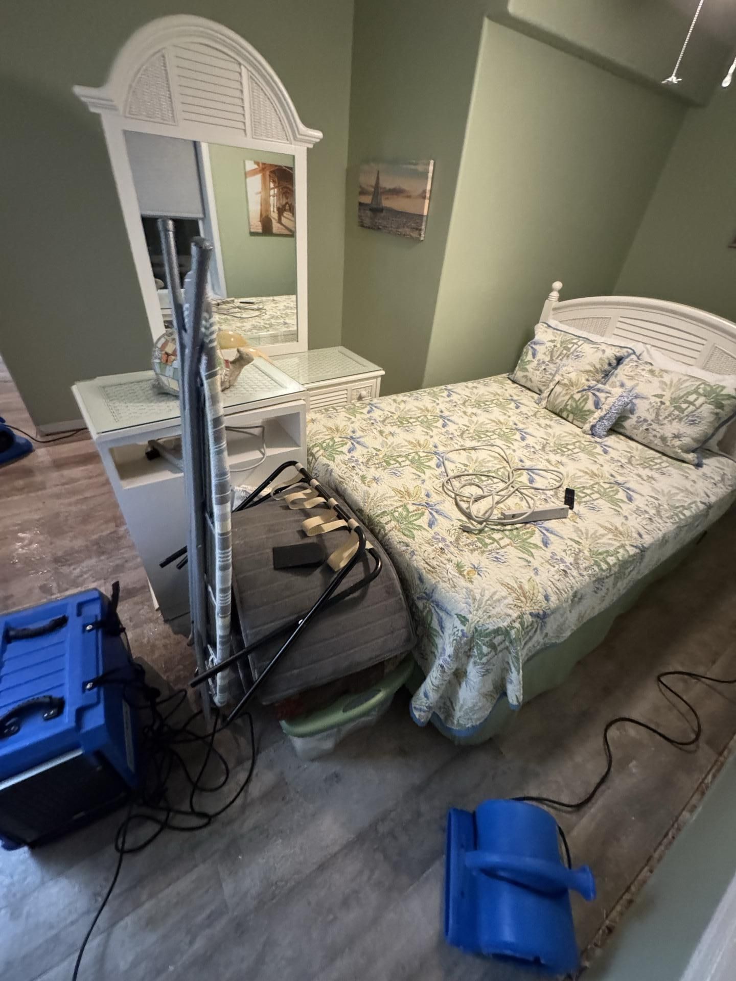 A bedroom with sage walls, a white bed, and a vanity, cluttered with an ironing board and two blue industrial air movers.