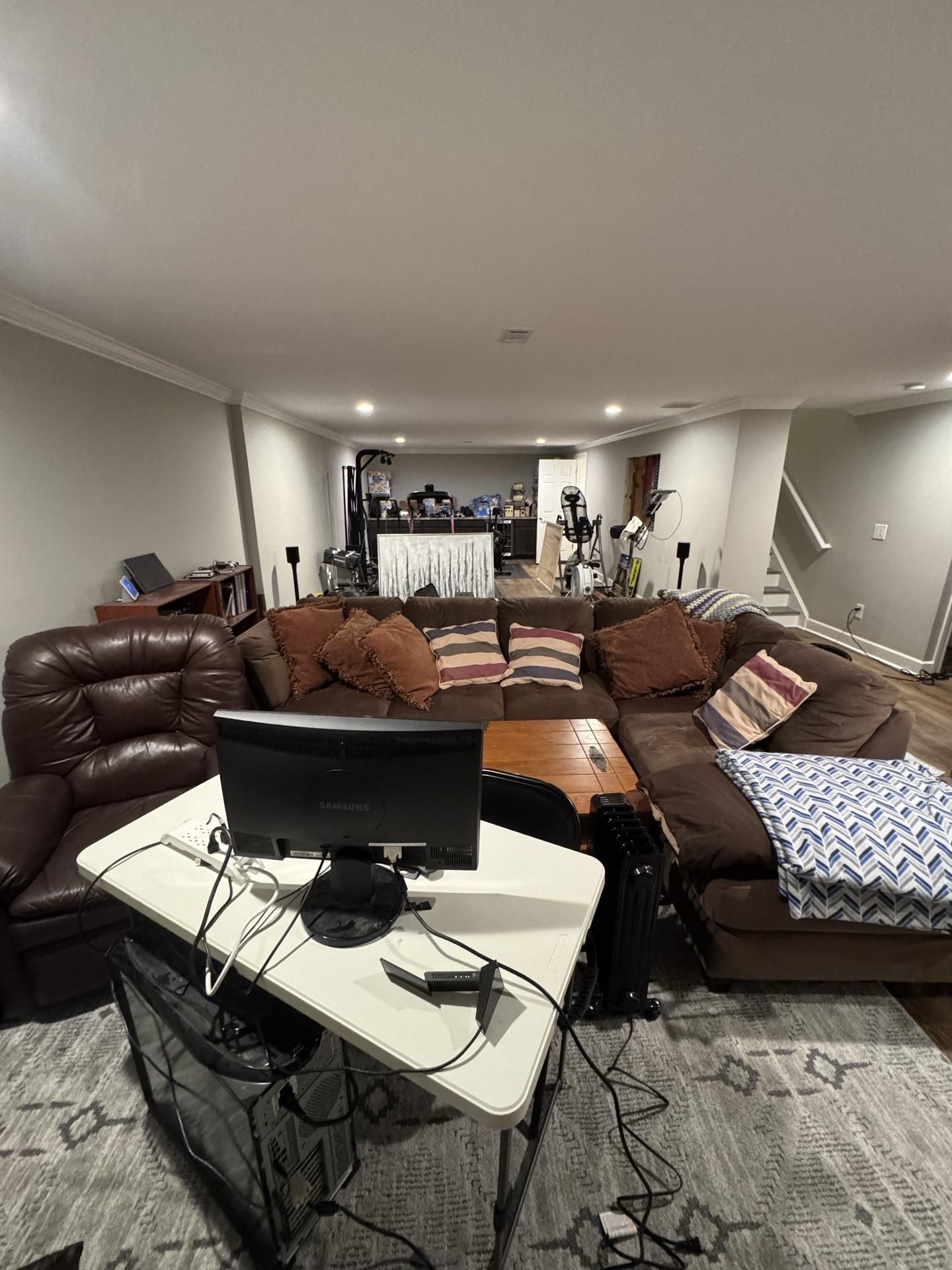A brown sectional sofa and leather recliner in a carpeted basement, with a small desk and monitor in the foreground.