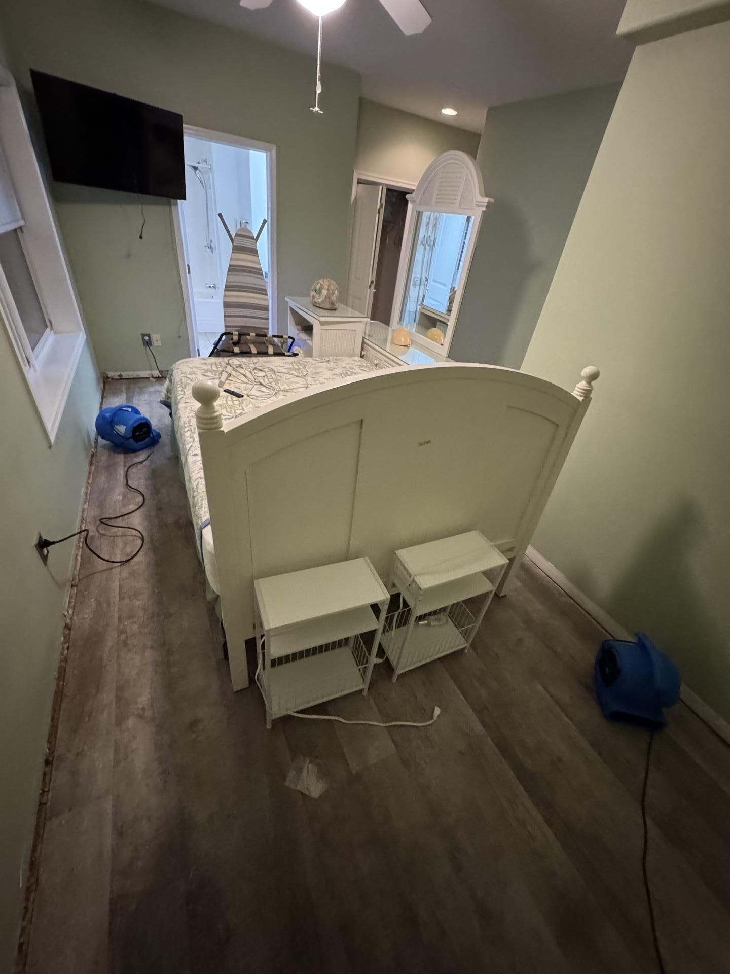 A bedroom undergoing water damage restoration with drying fans, a bed, and exposed flooring edges.