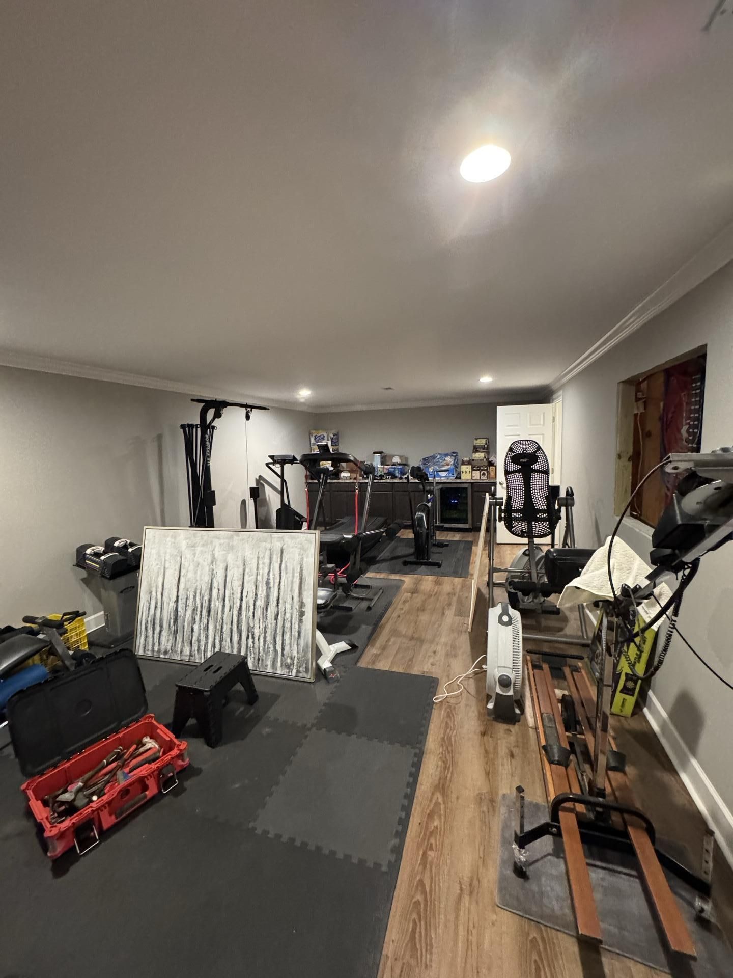 A home gym featuring workout machines, foam floor mats, a red toolbox, and a white privacy screen on a wood-look floor.