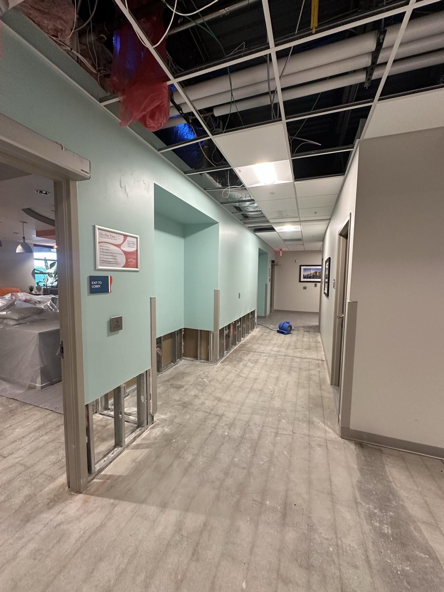 A hallway under construction with missing lower wall sections, exposed framing, and a partially open ceiling grid.