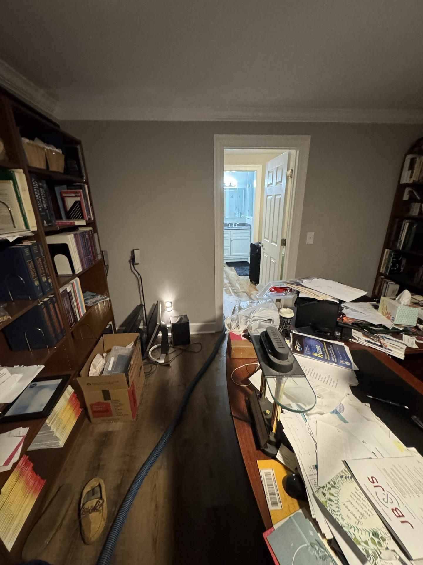 A cluttered home office with two tall wooden bookshelves, a desk covered in papers, and a doorway leading to another room.