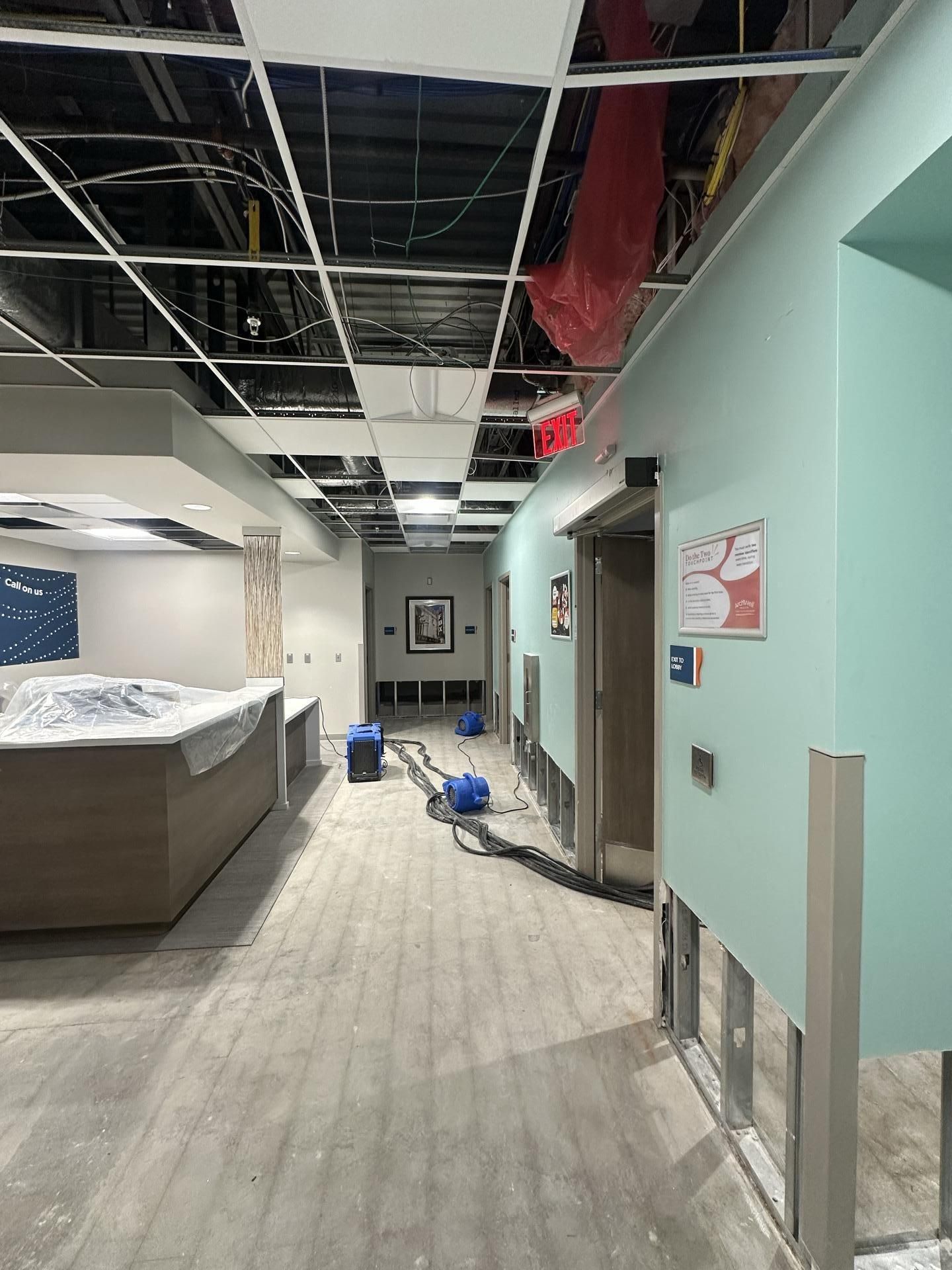 An interior office hallway under construction, featuring exposed ceiling grids, floor drying equipment, and bare walls.