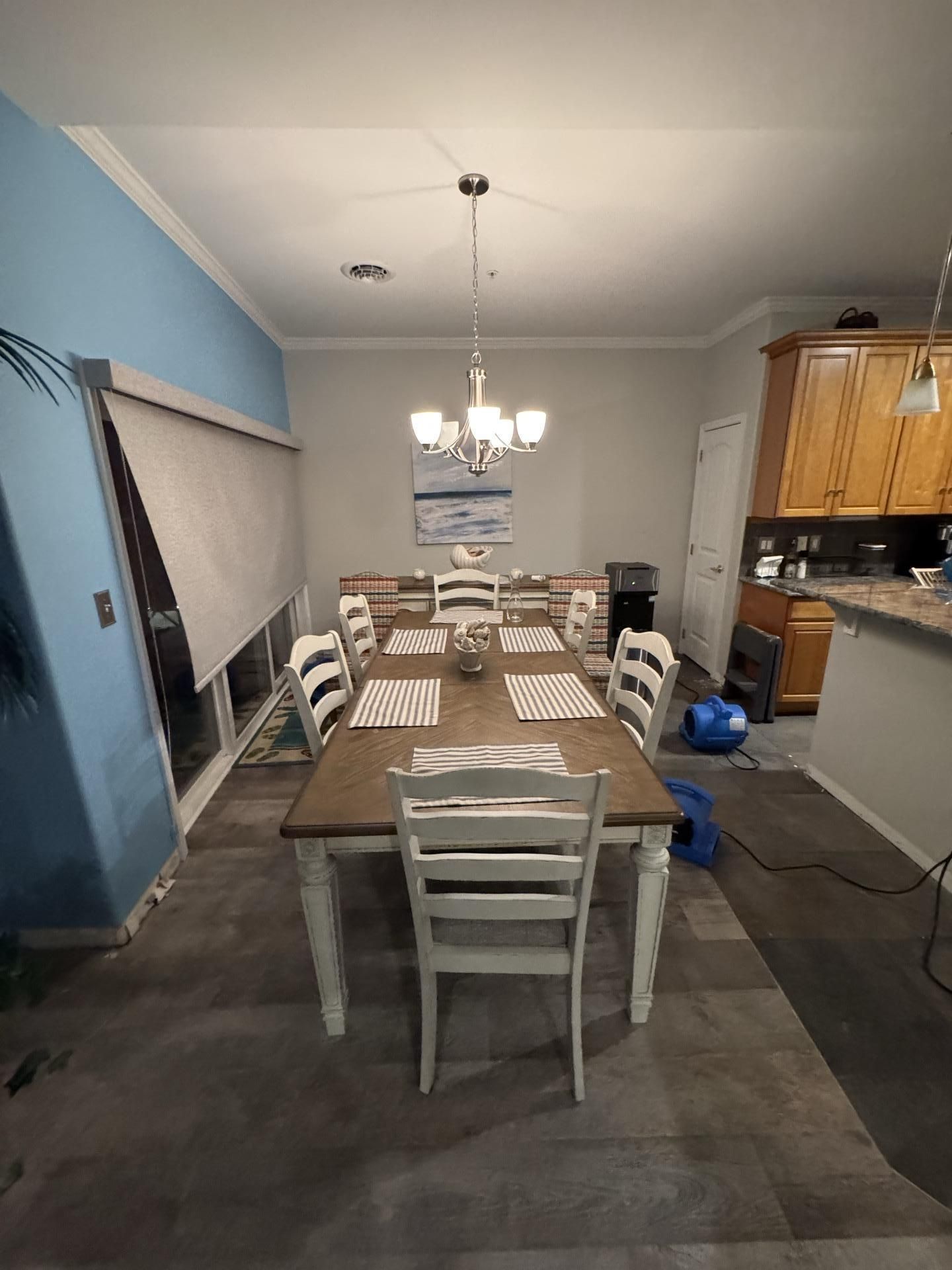 A dining room with a long wooden table, white chairs, a chandelier, and blue accent wall, with floor fans in the corner.
