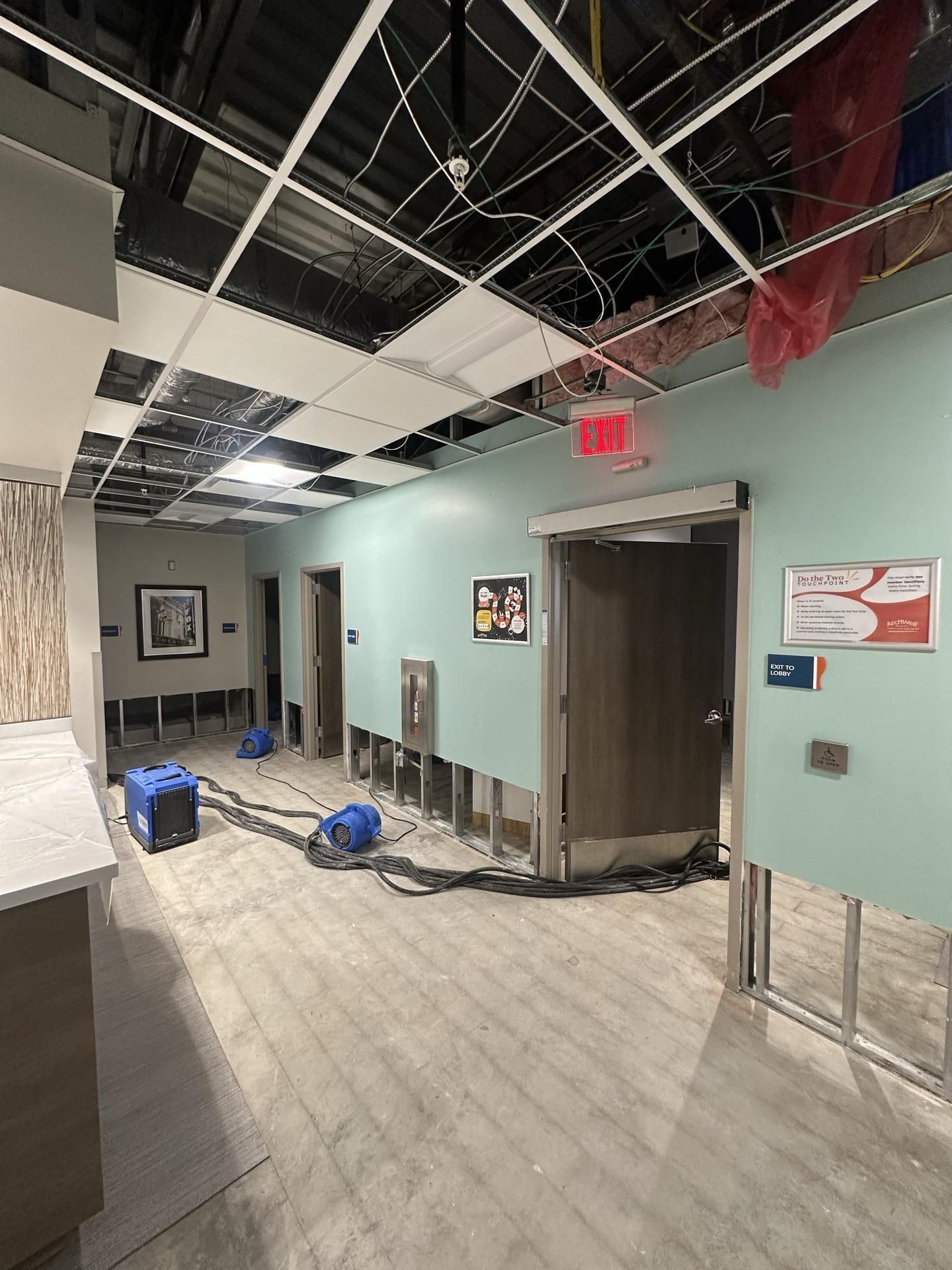 A hallway undergoing construction or renovation with exposed ceiling grids, partial wall panels removed, and air movers.