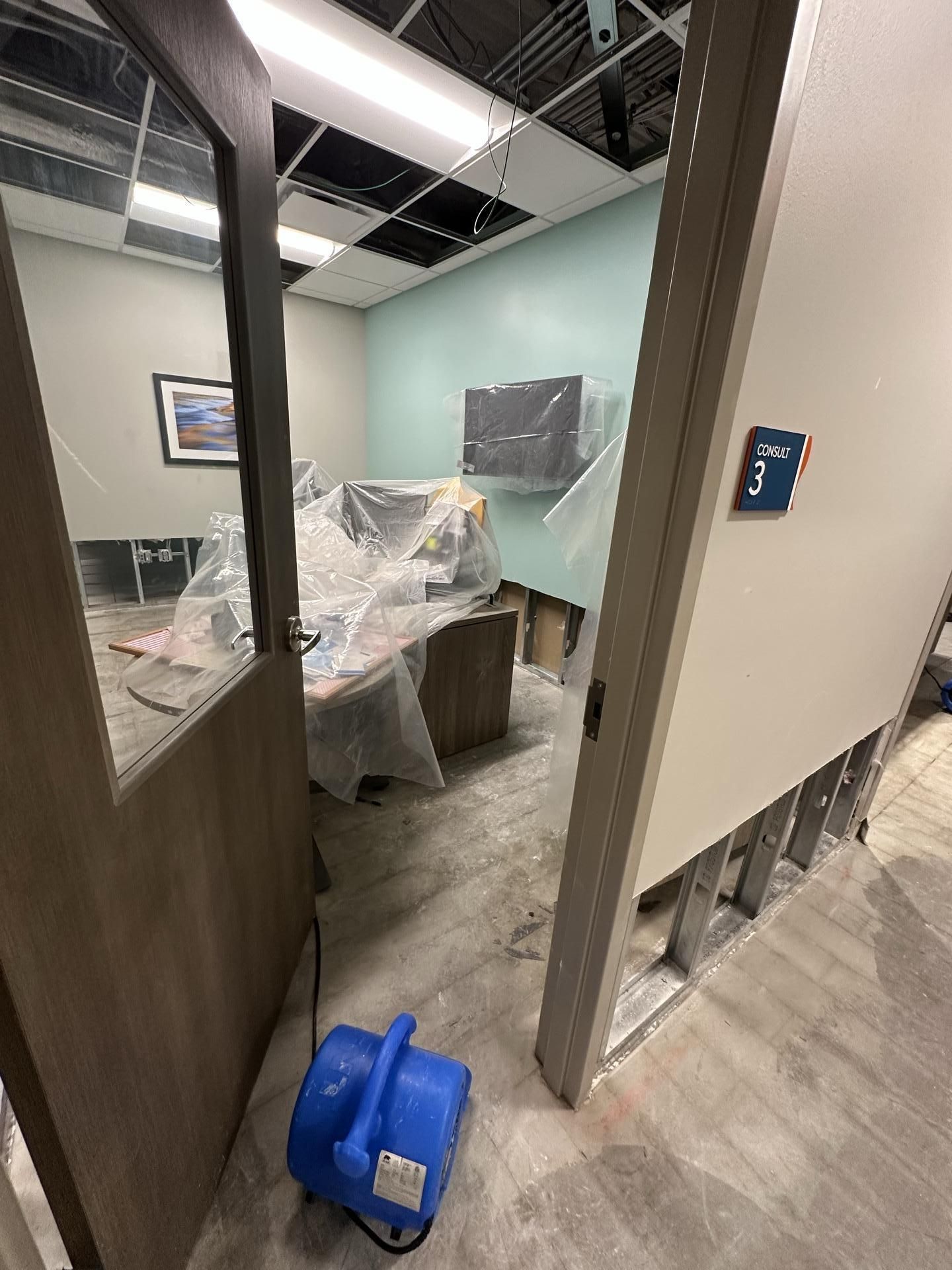 An office room undergoing restoration with plastic-covered furniture, an air mover on the floor, and open wall studs.