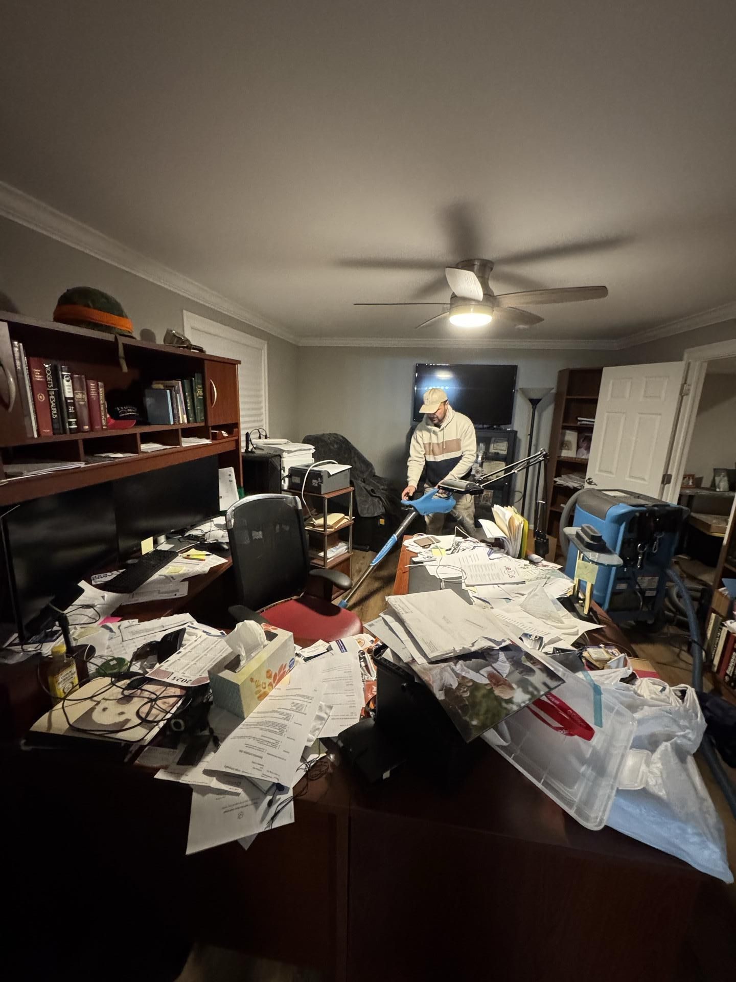 A person uses a blue carpet cleaning machine in a cluttered office with a desk covered in papers and a ceiling fan.