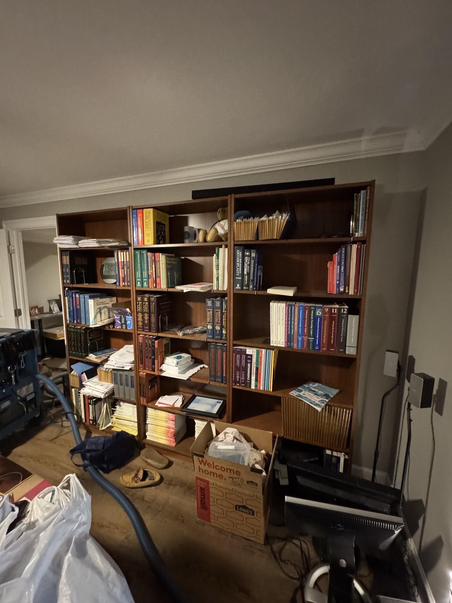 A tall, three-section wooden bookshelf filled with books, cluttered with boxes and cleaning equipment on the floor.