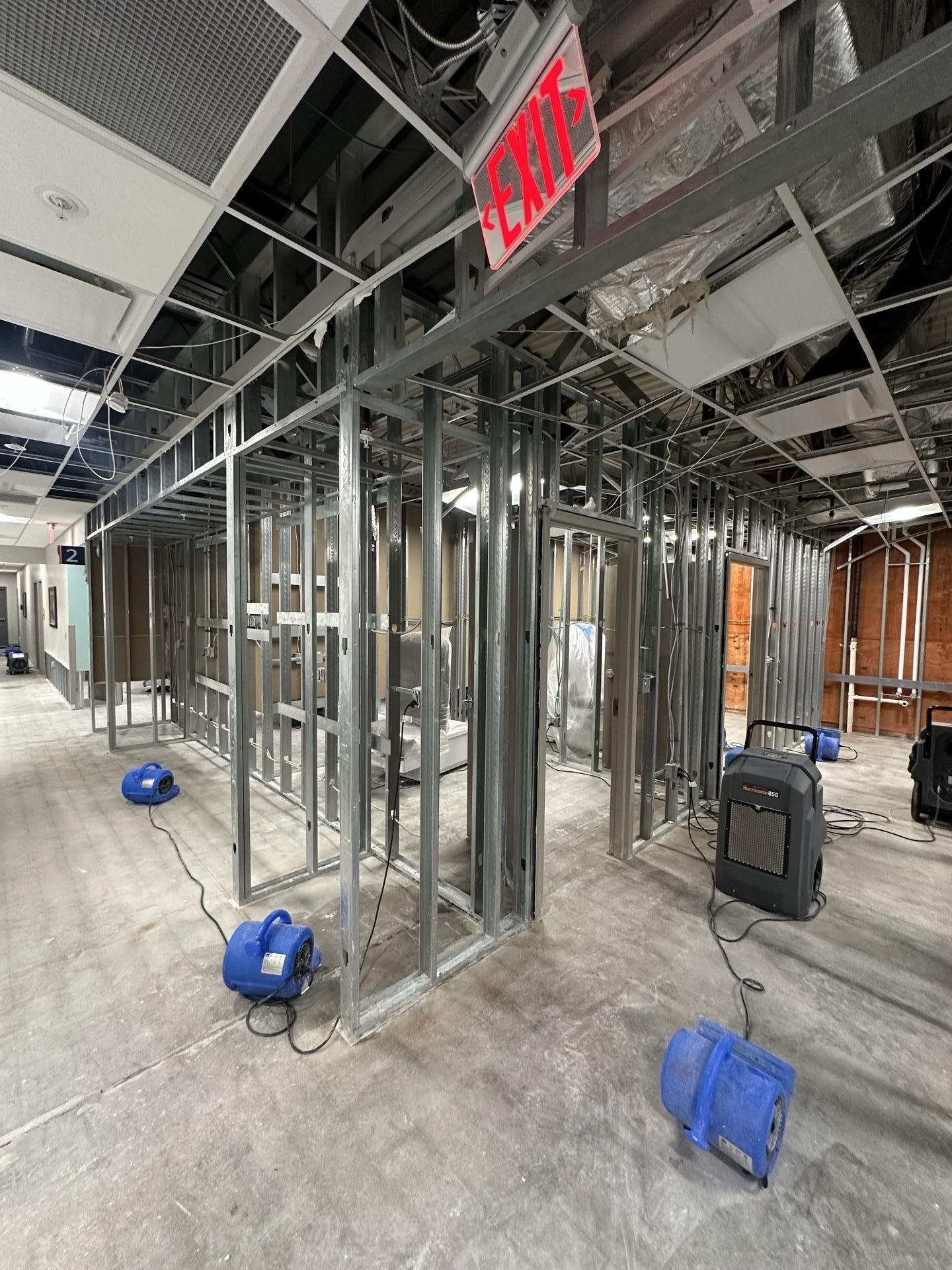 A construction site interior featuring metal wall framing, exposed ceilings, an exit sign, and blue air-drying fans.