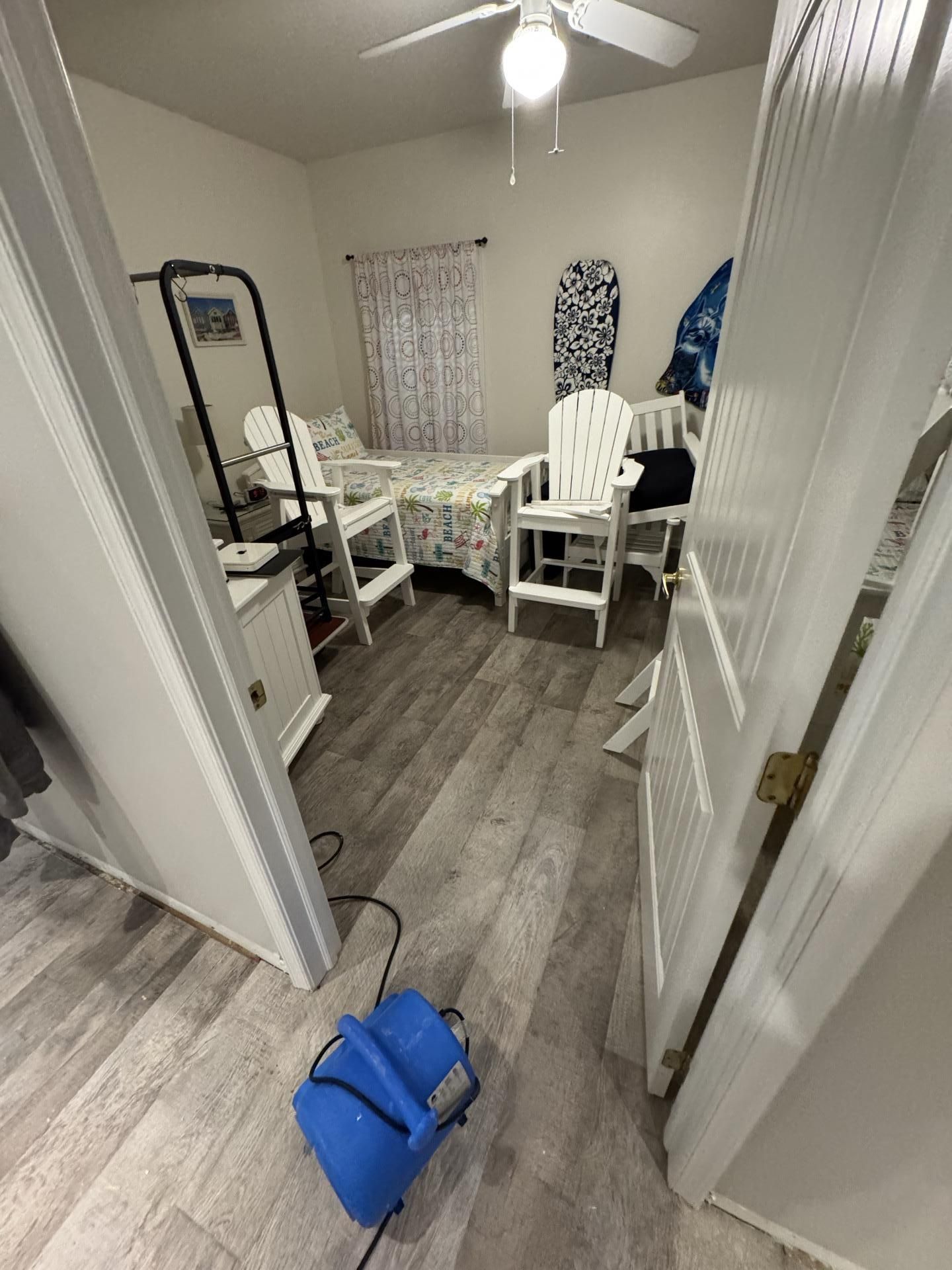 A bedroom with a blue floor fan, two white chairs, a bed with floral bedding, and surfboards against the back wall.
