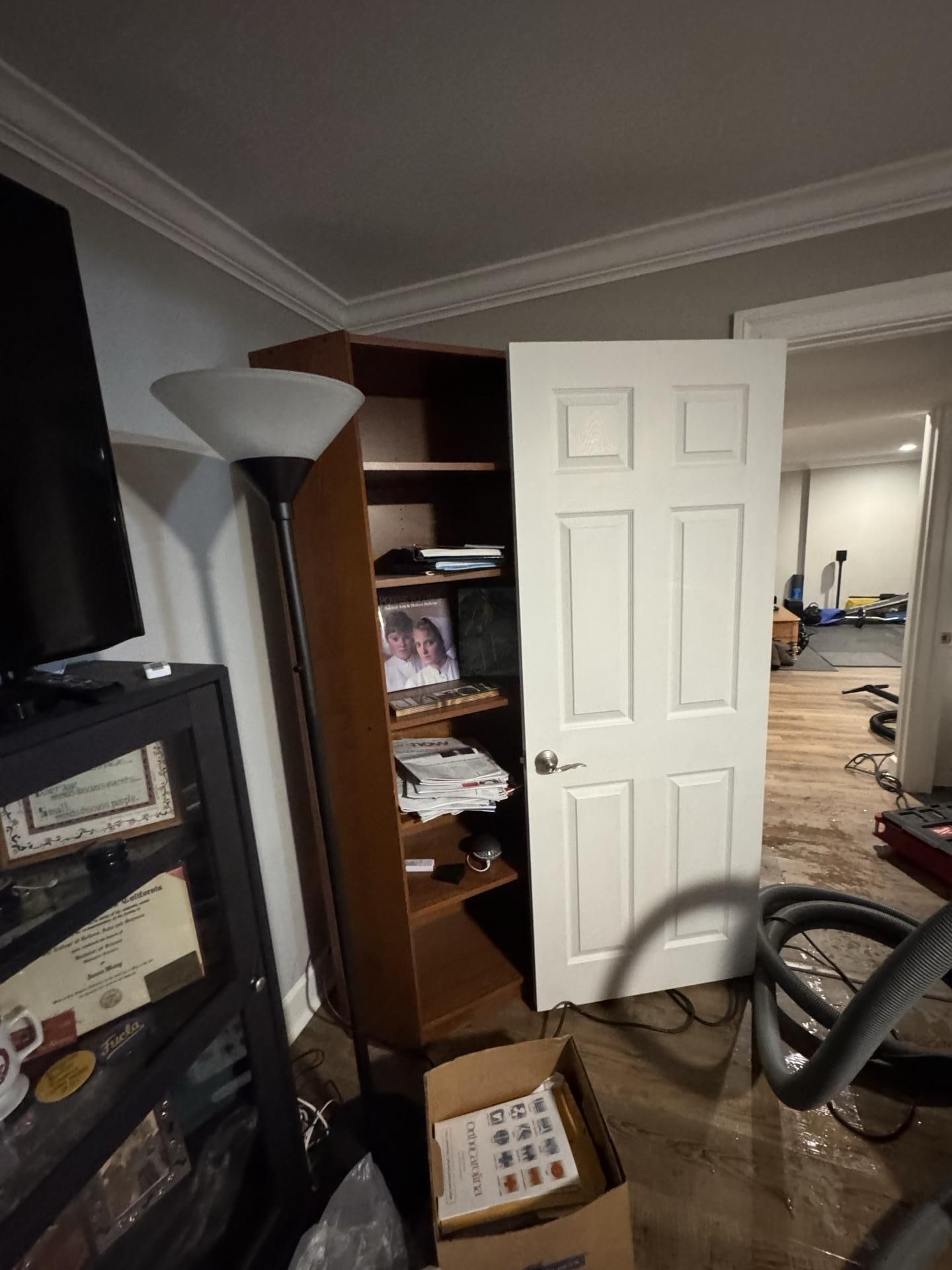 A room with a floor covered in water, a dark wood bookshelf, a floor lamp, a doorway, and a vacuum hose on the floor.