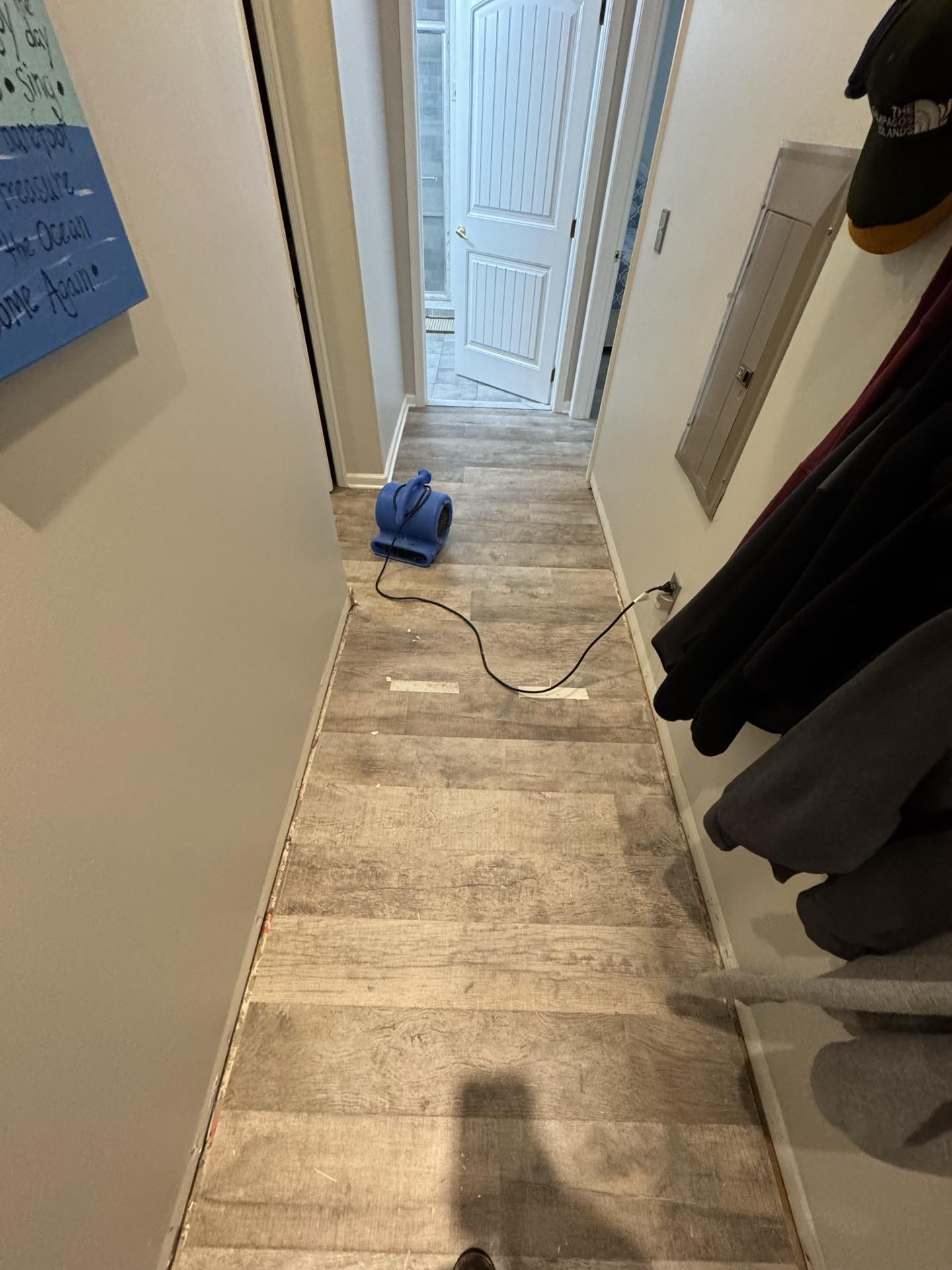 A blue industrial air mover sits on the wood-look flooring of a narrow indoor hallway, connected to a power outlet.