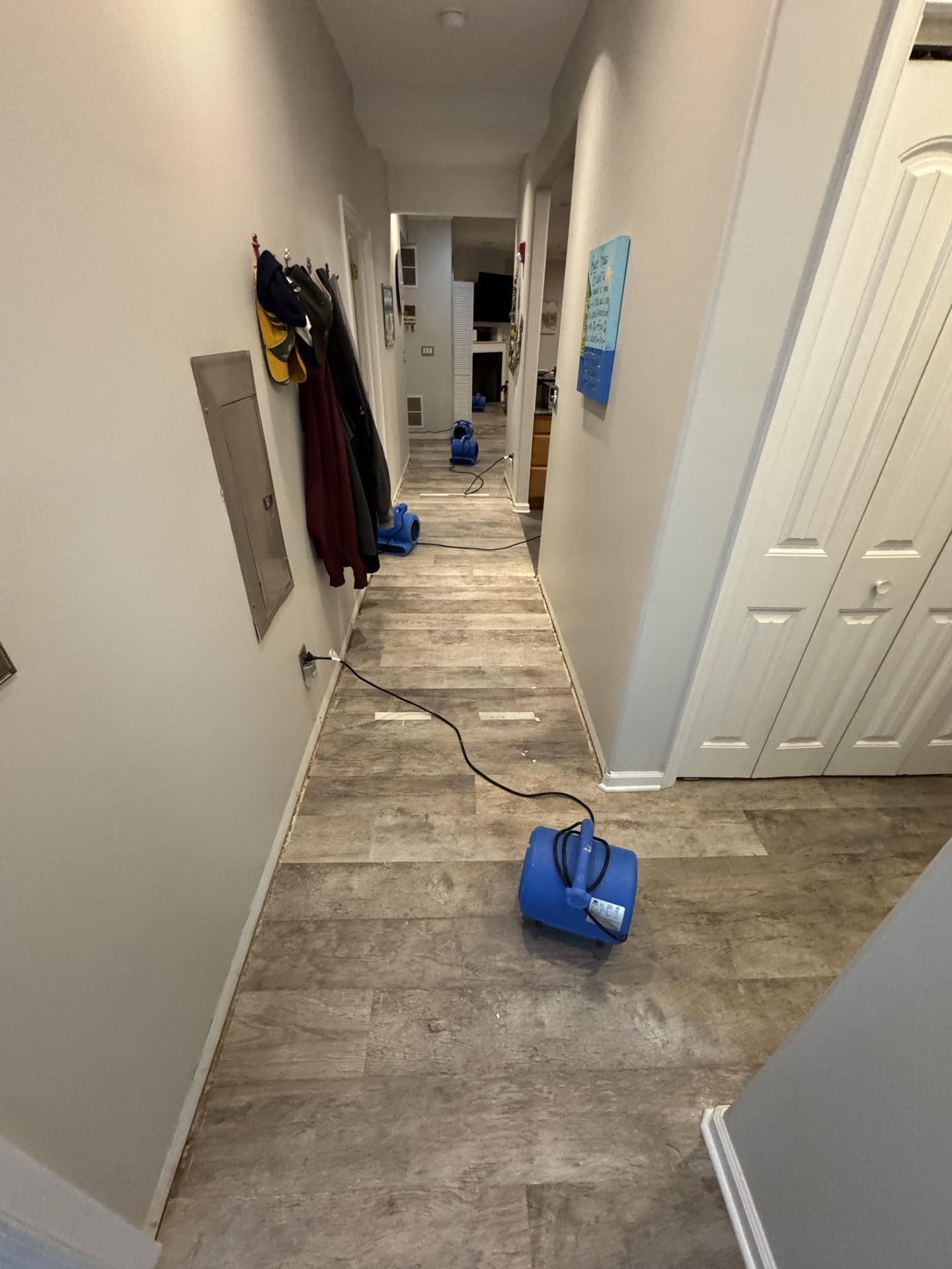 A hallway with a wooden floor, featuring two blue air movers positioned to dry the area, with coats hanging on the wall.