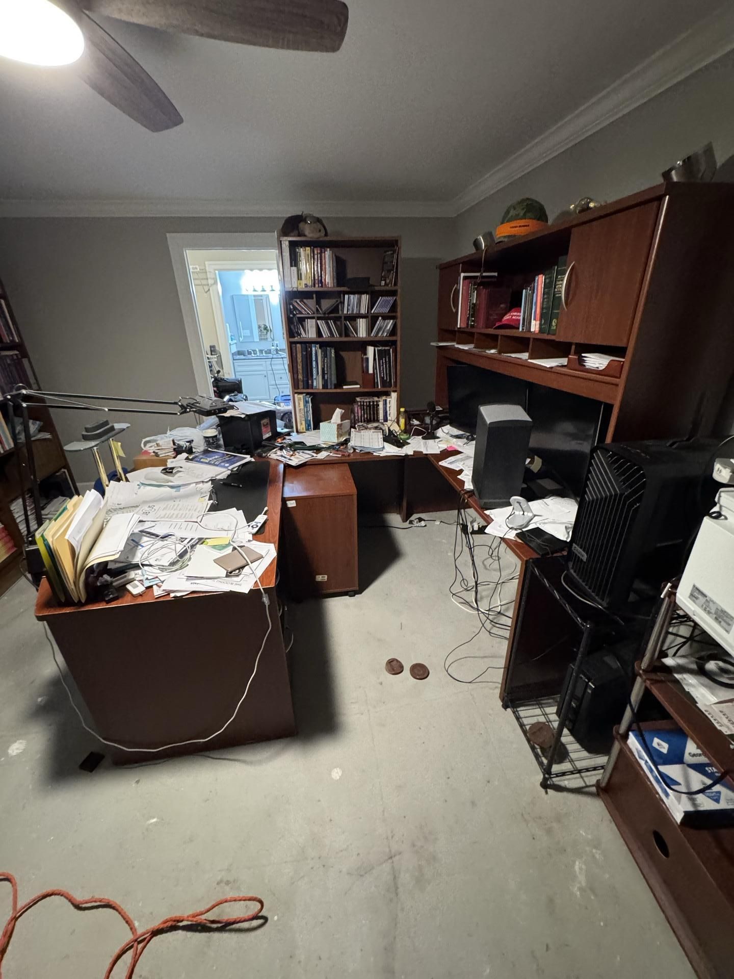 A cluttered home office with a dark wood L-shaped desk, a matching bookshelf, a desktop computer, and papers.