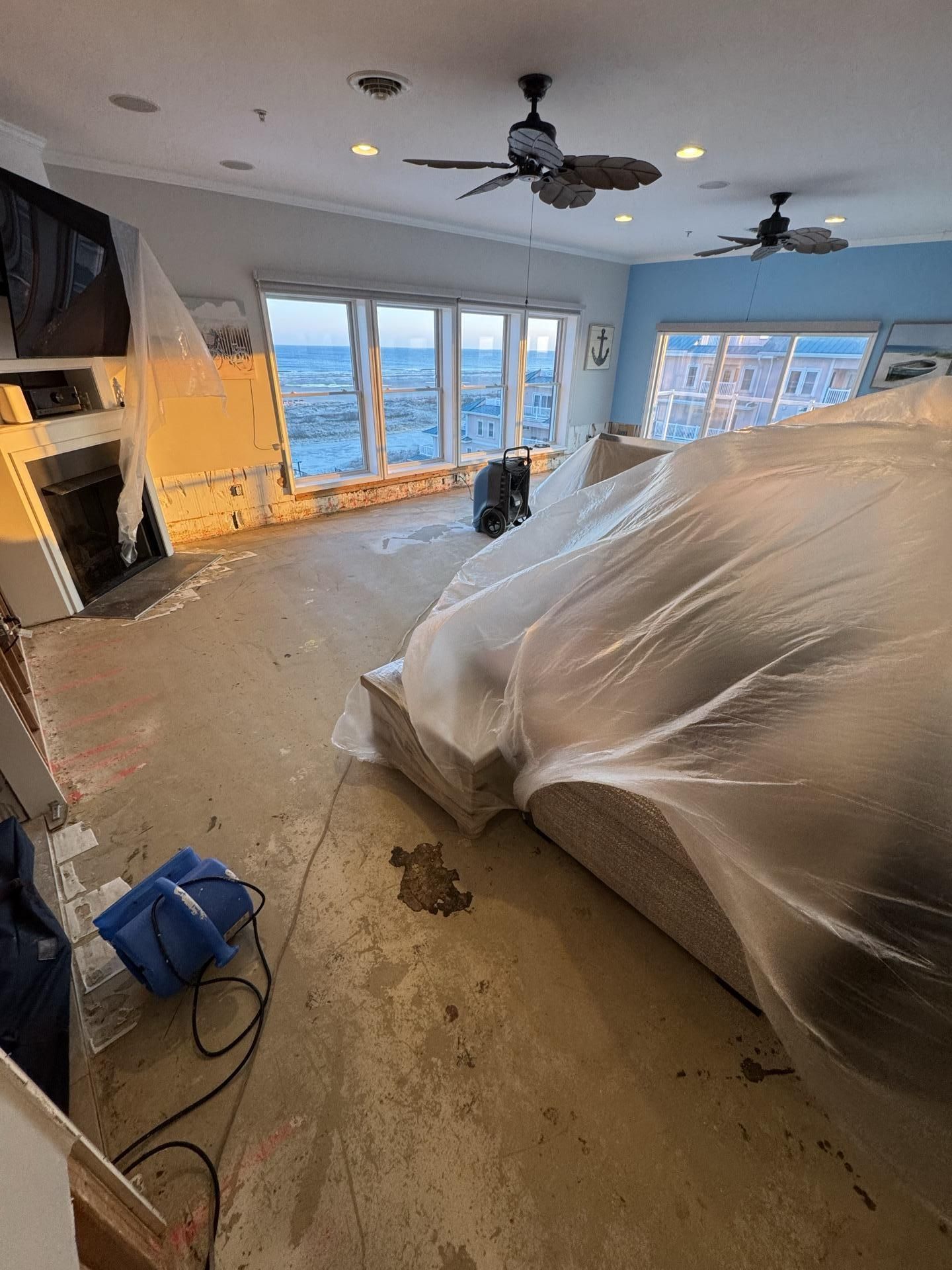 A room under renovation with exposed subflooring, plastic-covered furniture, and a blower fan on the floor.