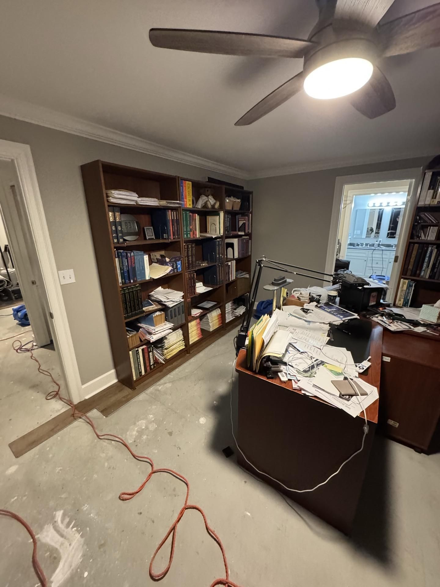 A cluttered home office features a large brown bookcase, a desk covered in papers, and a ceiling fan in a gray-walled room.