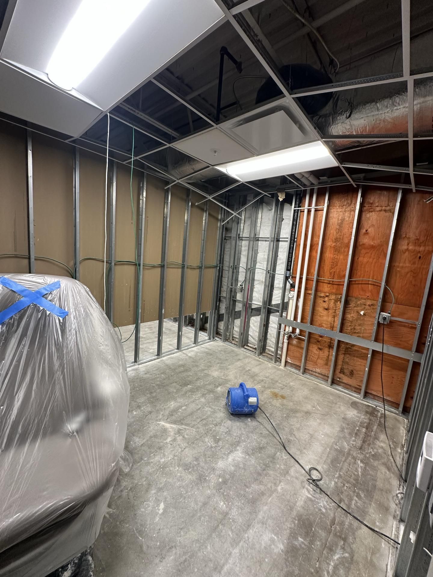 An interior room under construction with metal wall studs, exposed ceiling tiles, and a small blue fan on the concrete floor.