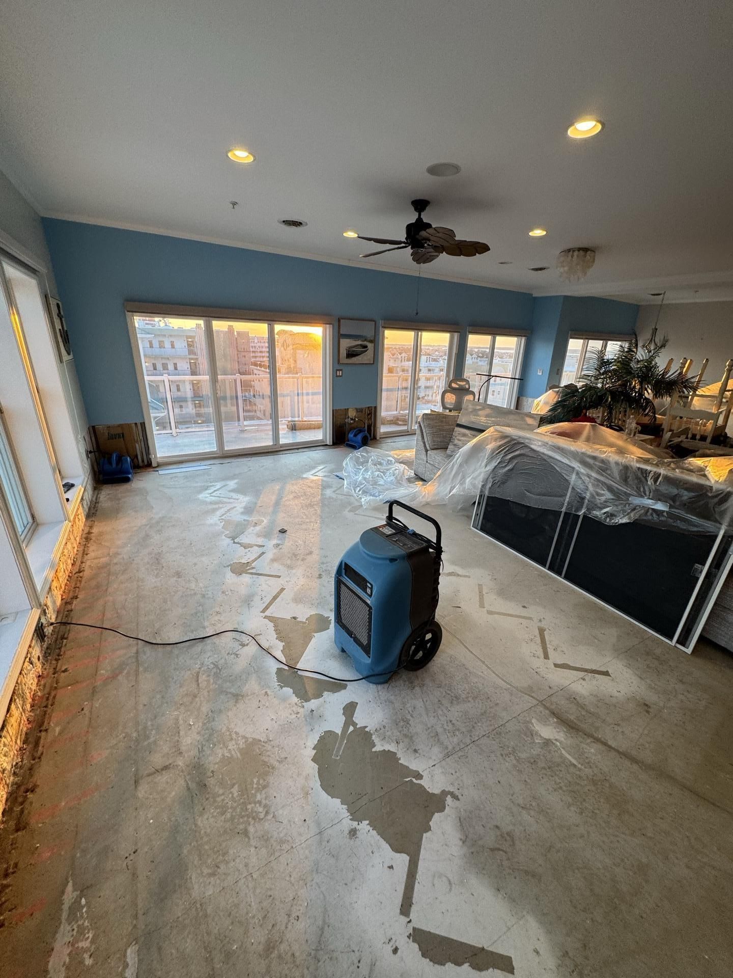 A blue dehumidifier sits in a room with stripped floors and blue walls, undergoing water damage restoration.