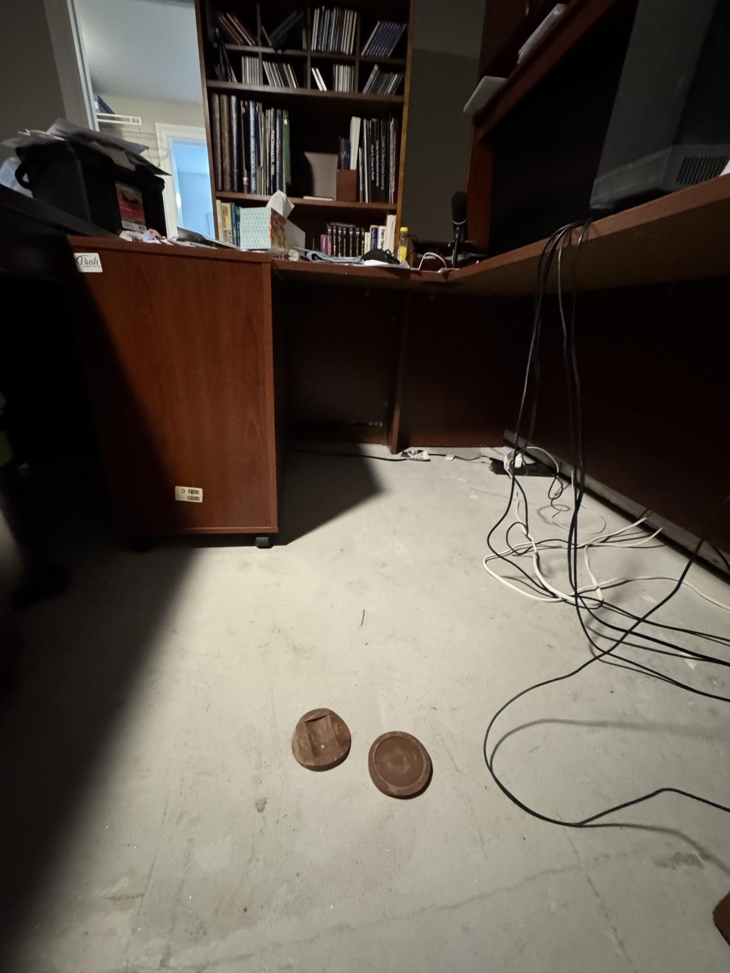 A low-angle view of a home office floor featuring a large wooden desk, shelving, tangled black cables, and two coasters.