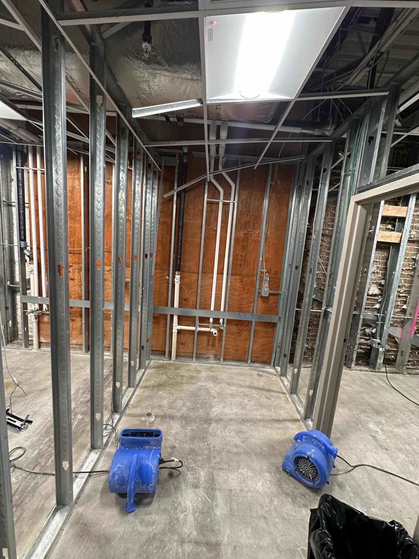 Framed wall studs and exposed plumbing in an indoor construction site with two small blue floor fans on the concrete floor.