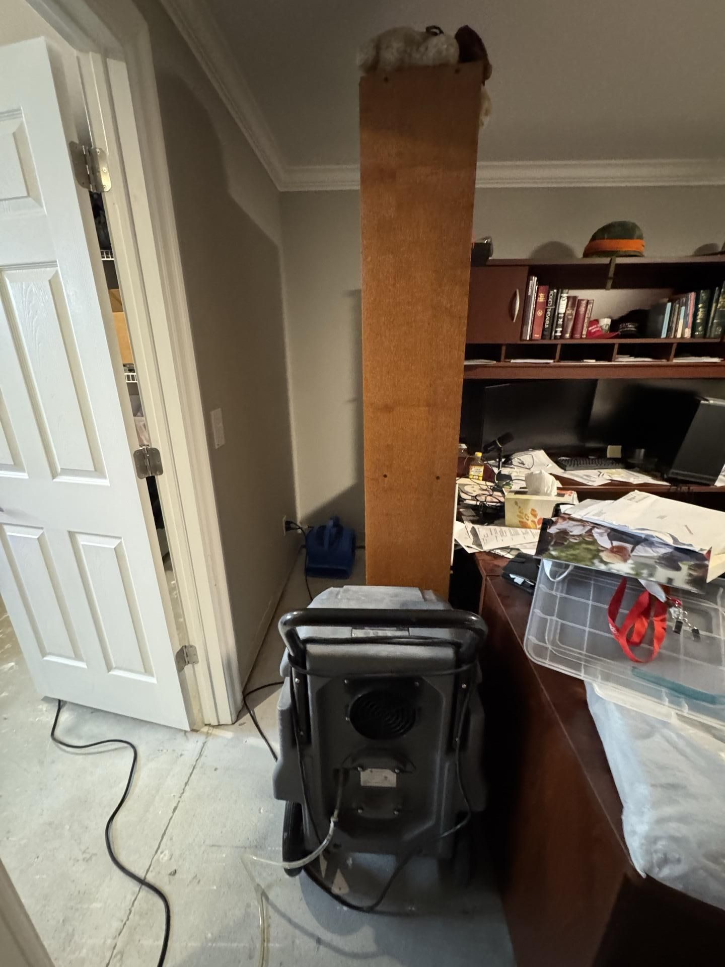 A gray industrial dehumidifier sits on a floor near a doorway and a desk in a room being treated for water damage.
