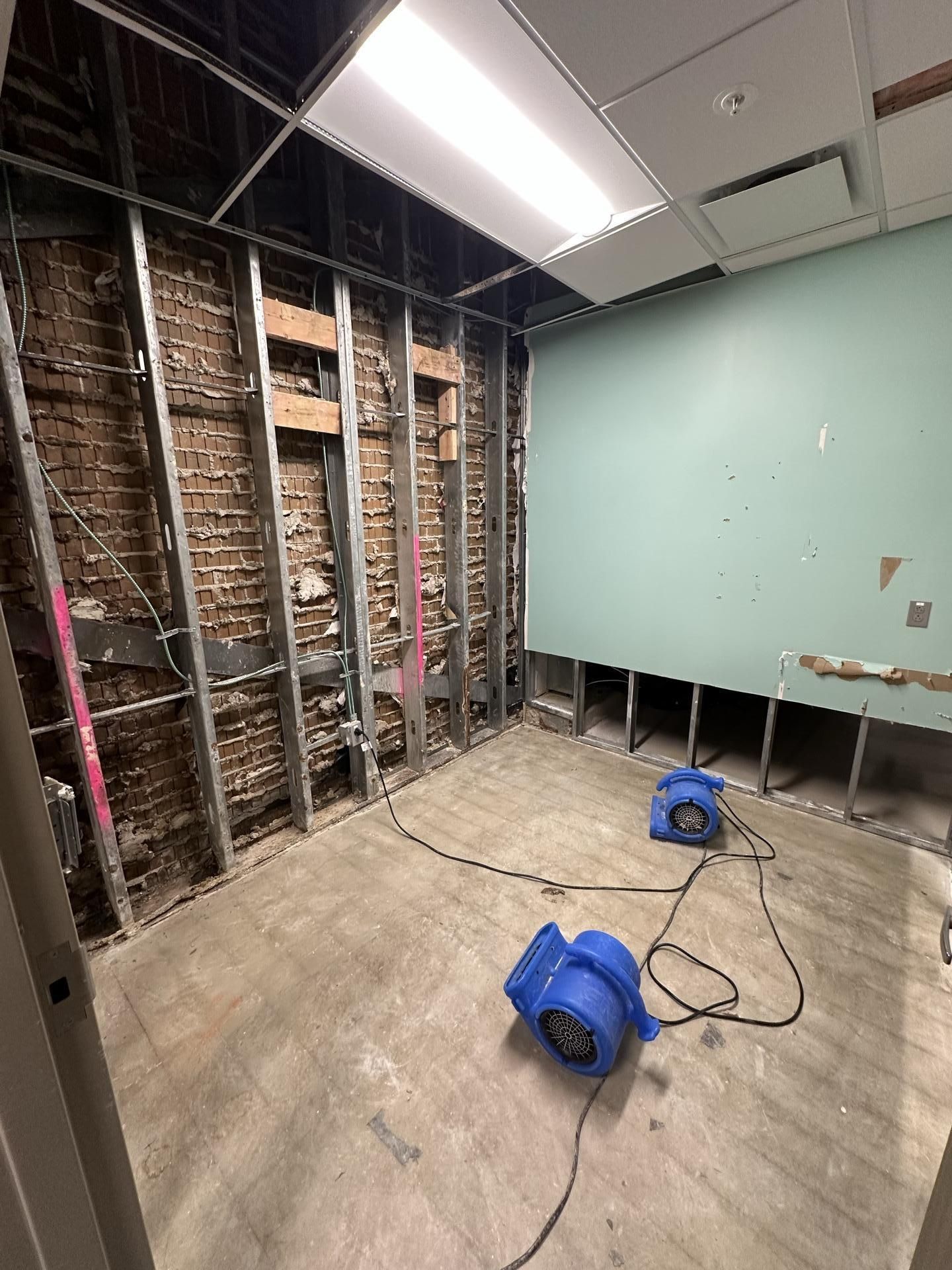 Interior of a room under construction with exposed brick walls, metal framing, drywall, and two blue floor drying fans.