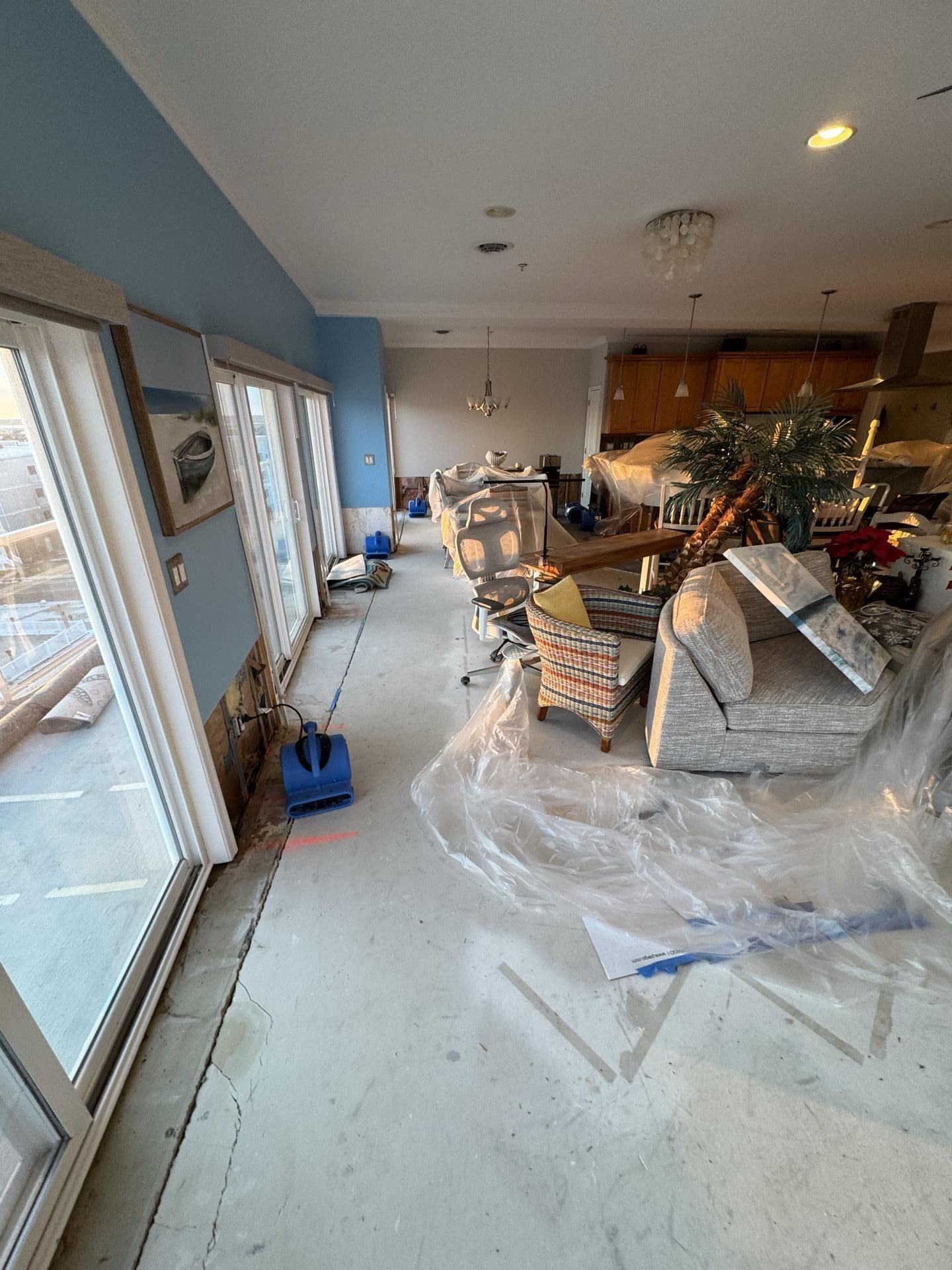 A home living area undergoing water damage restoration, with bare concrete floors, a drying fan, and furniture under plastic.